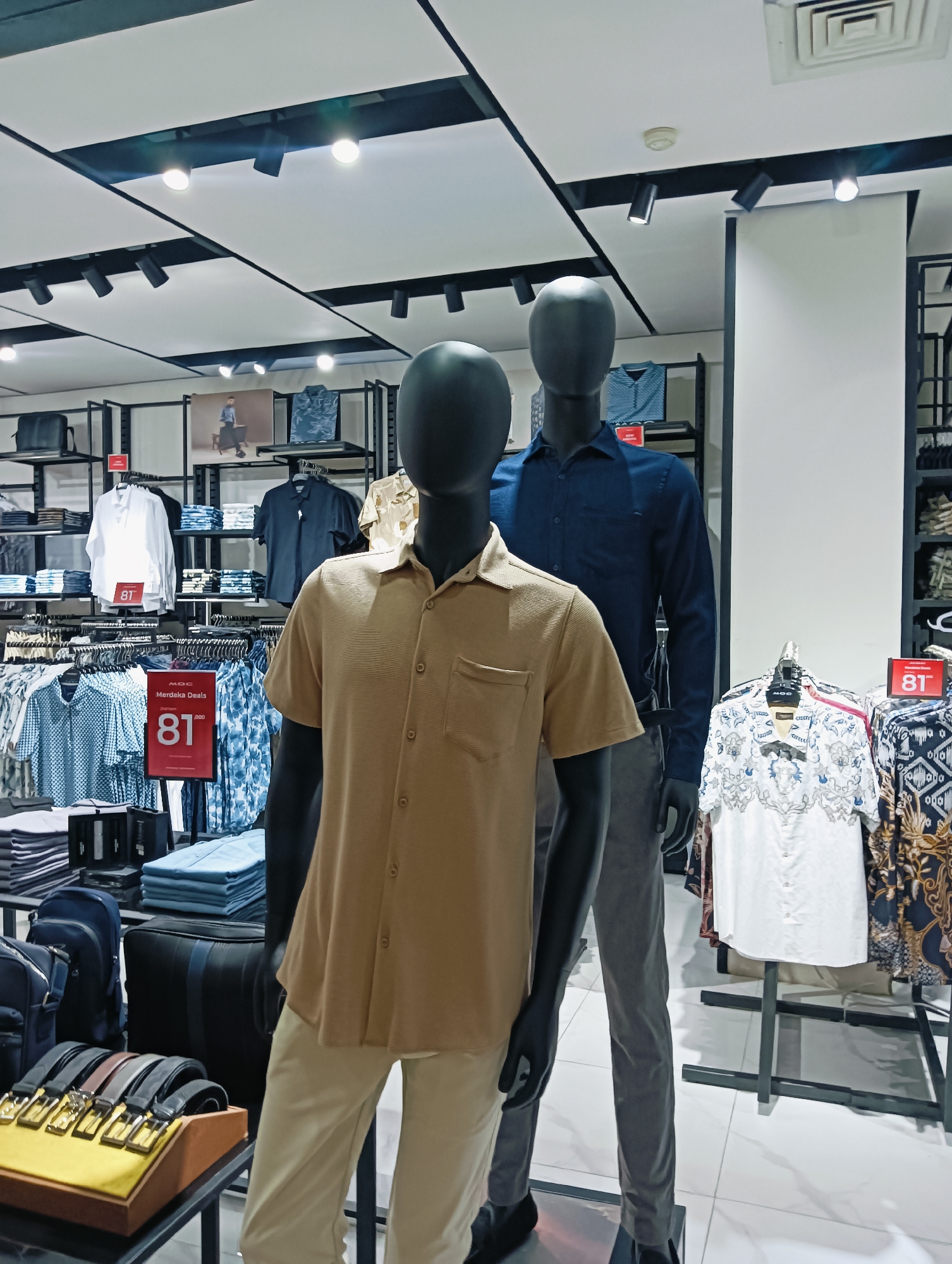 The image depicts a retail clothing store, likely a men's fashion store, located in Batam, Indonesia. Two male mannequins are prominently displayed in the foreground. The mannequin on the left is dressed in a short-sleeved, light brown collared shirt and light-colored pants. The mannequin on the right wears a long-sleeved, dark blue collared shirt and grey pants. Behind the mannequins, racks of clothing are visible, including various shirts with different patterns and colors. Some items are displayed folded on shelves. A red sign with the number "81" is visible, suggesting a sale or promotion. The store features modern lighting with recessed spotlights and track lighting. Shelves and racks are made of dark metal, and the floor is tiled in a light color. Bags and belts are also displayed on lower shelves. The overall environment suggests a clean, well-organized retail space.
