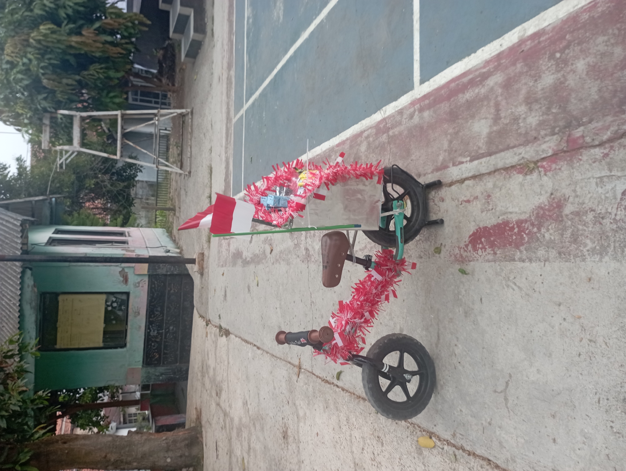 A balance bike decorated with red and white tinsel and an Indonesian flag is parked on a concrete pathway next to a blue and white court, possibly a basketball or tennis court. Behind the bike, a small, light green building with a corrugated metal roof stands under a tree. A metal ladder structure is visible to the right of the building, and a set of dark steps is to the far right. The environment suggests an outdoor area, likely in a school or public space in Purwakarta, Indonesia, given the patriotic decorations. There are no people visible in the image.