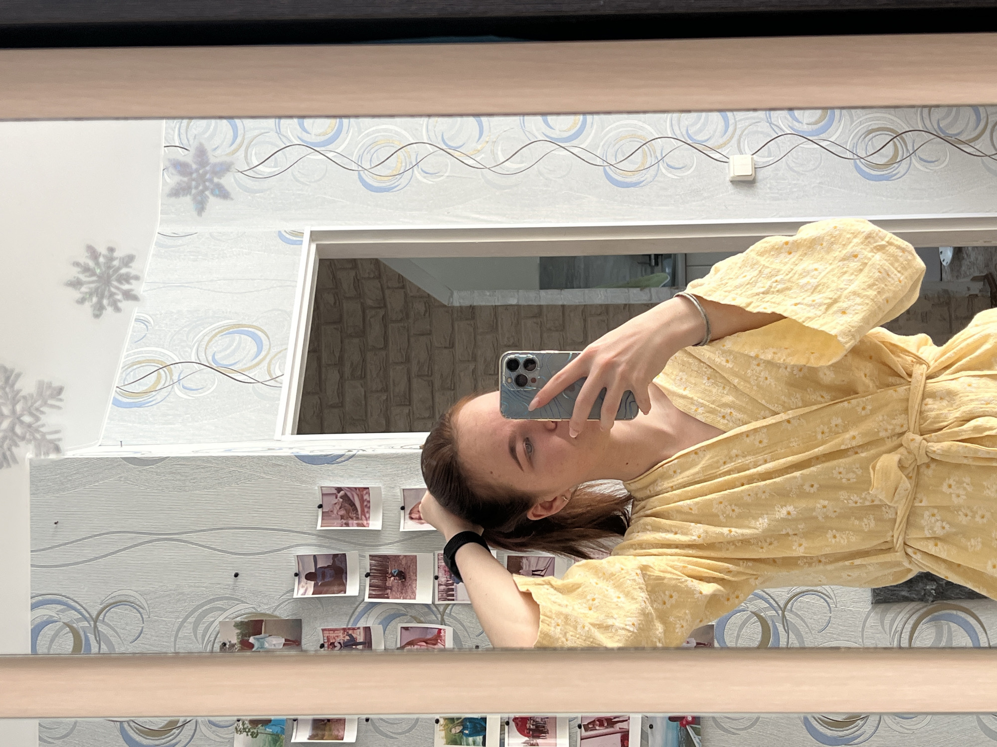 An indoor mirror selfie taken in an apartment in Chaikovsky, Russia, features a young female subject, visible from the chest up. She wears a yellow wrap-around robe adorned with a white floral pattern and secured by a tied belt. Her left hand holds a light blue smartphone, partially obscuring the lower half of her face, while her right arm is raised, with her hand positioned near the back of her head, appearing to adjust her light brown hair, which is pulled into a ponytail. A black hair tie is on her right wrist, and a silver-toned bracelet is on her left. The background, visible in the mirror's reflection, consists of a wall covered in patterned wallpaper featuring light blue, grey, and gold circular and wavy line motifs. Numerous small, rectangular photographs are affixed to the wallpaper on the left side of the reflection. The ceiling displays two reflective, snowflake-shaped decorations. A doorway leading to an adjacent room with a light-colored, textured wall is visible in the background reflection. A white electrical wall switch with a visible wire extends on the right side of the reflection. The mirror is framed by light-colored wood on the left and a darker material on the right.
