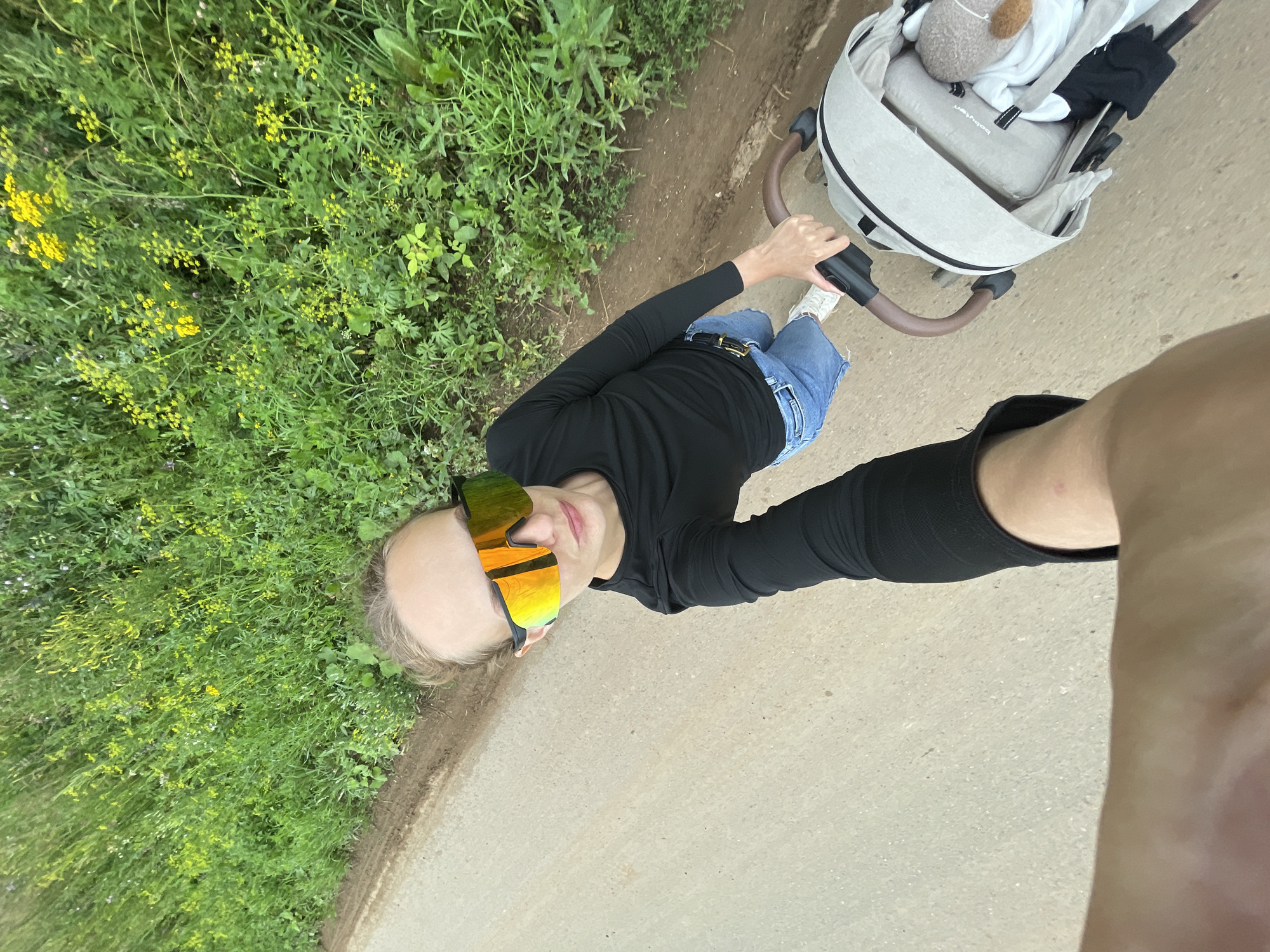 The image shows a person pushing a stroller along a dirt path next to a field of green vegetation with yellow wildflowers. The person is wearing a black long-sleeved shirt, denim shorts, and white sneakers. They are also wearing large, reflective sunglasses with orange and yellow lenses. The stroller is light gray with a brown handle and appears to contain a stuffed animal. The perspective is from the person's point of view, as they are holding the camera in their outstretched arm. The scene appears to be outdoors in a rural or semi-rural setting, possibly on the outskirts of the village of Podshivalovo, Russia, given the context.