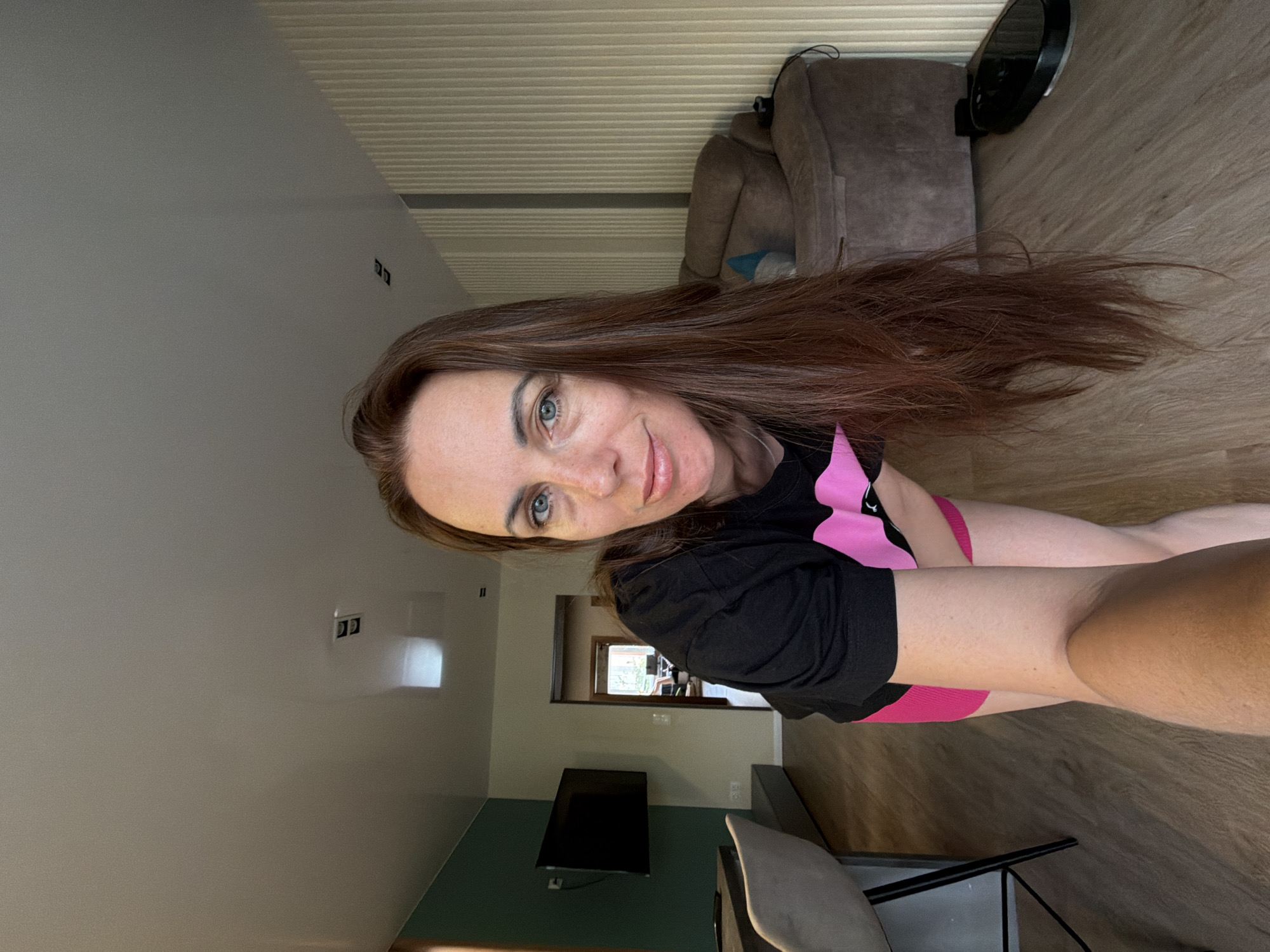 A young woman with long, reddish-brown hair and blue eyes is pictured indoors in what appears to be a residential setting in Birobidzhan, Russia. She is looking directly at the camera with a slight smile, suggesting she is taking a selfie. She is wearing a black t-shirt with a pink graphic design and pink shorts or leggings, and is positioned on the floor with her legs bent or extended.
The scene is a well-lit room with light-colored walls and a section painted in a teal or dark green hue. The floor is made of wood or laminate. In the background, a brown upholstered armchair or sofa is visible, along with a dark, circular object that might be a robot vacuum cleaner or a fan. A wall-mounted television is present on the teal wall. A doorway in the mid-background reveals another room with natural light coming through a window. A partial view of a chair with a gray seat is seen in the foreground. The ceiling has a horizontally corrugated or textured appearance. The overall impression is of a casual, relaxed moment captured during the daytime. No specific text is clearly visible on clothing or in the environment.