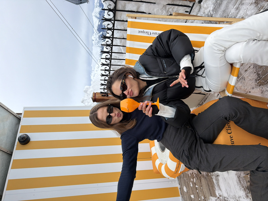 The image captures two young women posing for a photo in an outdoor, snowy, mountain setting, likely a ski resort or a high-altitude lounge area. The overall impression is one of a relaxed, fun social occasion.
In the foreground, to the left, is one woman with long, straight brown hair, wearing dark, rectangular sunglasses, a dark long-sleeved top, and dark pants. She is holding an orange-tinted, uniquely shaped glass, possibly a champagne flute, with a pointed base. Her lips are slightly pursed in a playful pout as she looks towards the camera. She appears to be sitting or leaning on an inflatable, round orange and white striped cushion or float, which has the brand "Veuve Clicquot" printed on it in black lettering.
Behind and slightly to the right of her is the second woman. She has shorter, light brown hair, possibly styled up or pulled back, and is wearing brown-patterned sunglasses and white earbuds. She is dressed in a black puffer jacket over a white long-sleeved top and white pants. Her right hand is raised, making a gesture that resembles a peace sign or a stylized 'V'. She is seated on a wooden deck chair with yellow and white striped fabric, which also prominently displays the "Veuve Clicquot" brand name.
The background shows a stark, snow-covered mountain landscape under a bright, cloudy sky. A black ornate metal fence runs along the edge of what appears to be a viewing platform or balcony, with significant snow accumulation beyond it. Thin wires, possibly from a ski lift or gondola, stretch across the upper part of the frame against the sky. To the far left, a yellow and white striped wall, matching the branding elements, is visible, featuring a small black dome-shaped fixture, possibly a camera or light. The ground is a wooden deck, partially covered in scattered snow. The bright lighting suggests it is daytime. The women appear to be enjoying themselves and are dressed for cold weather.