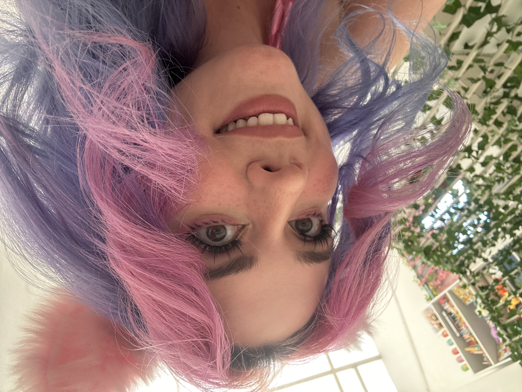 A woman with pink and purple hair and cat ears is smiling at the camera. She is indoors, with a window and shelves of items visible in the background. A latticework structure covered in greenery can also be seen. The location is possibly City of Zagreb, Croatia.