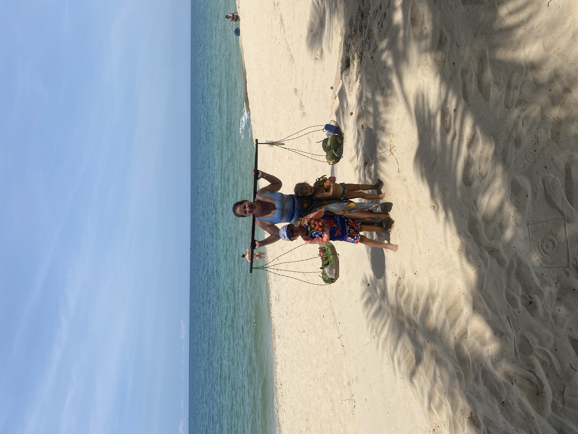 A woman carries a yoke with baskets of fruit on her shoulders on a sandy beach in Phu Thuy Ward, Vietnam. She is wearing a blue tank top and a patterned skirt. Two children stand beside her. A boy on her left, wearing a blue and orange swim shirt and patterned swim trunks, holds a bunch of red fruit. A younger child on her right, wearing only a diaper, holds a few oranges. In the background, the turquoise ocean meets a clear blue sky. Two people are visible in the water, and another person sits on the sand in the distance. Shadows of palm fronds are cast across the sand.