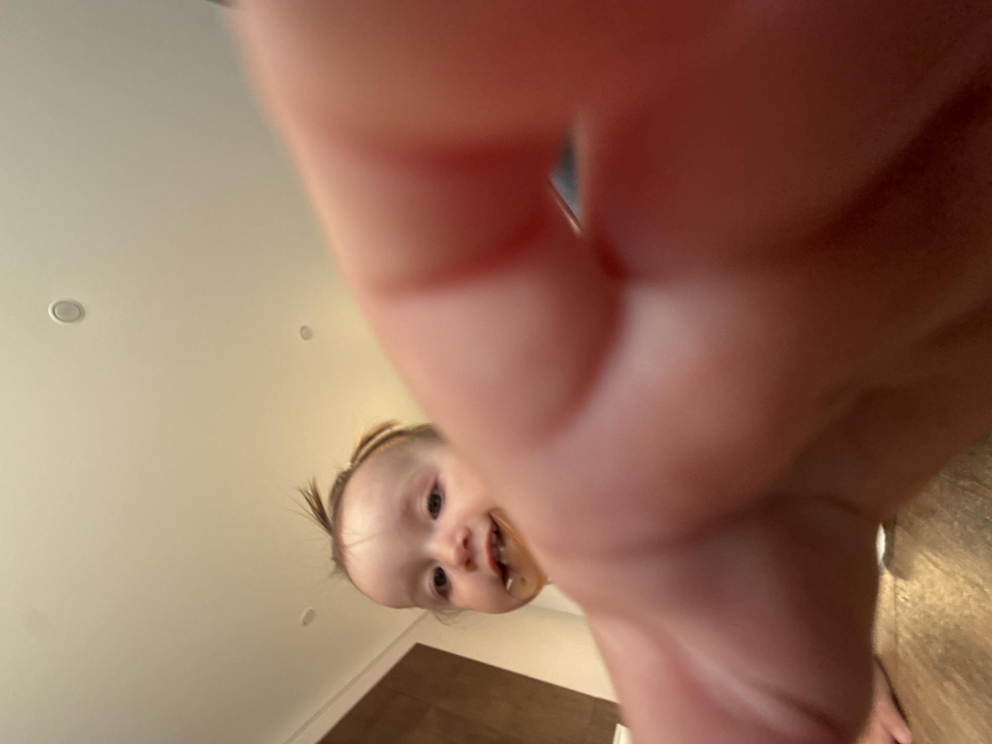 The image depicts a close-up, low-angle shot of a young child, likely a toddler, with a pacifier in their mouth. The child's face is the primary subject, with their eyes looking towards the camera. A blurry hand, presumably belonging to the person taking the photo, occupies the foreground, obscuring a significant portion of the frame. The background appears to be an indoor environment, possibly a room with a wooden floor and a cabinet or furniture. The ceiling is visible in the upper portion of the image, with recessed lighting fixtures. The overall impression is that of a candid, informal photograph taken by someone very close to the child. There is no information available to determine the location of this scene in the city of Подшивалово, Russia.