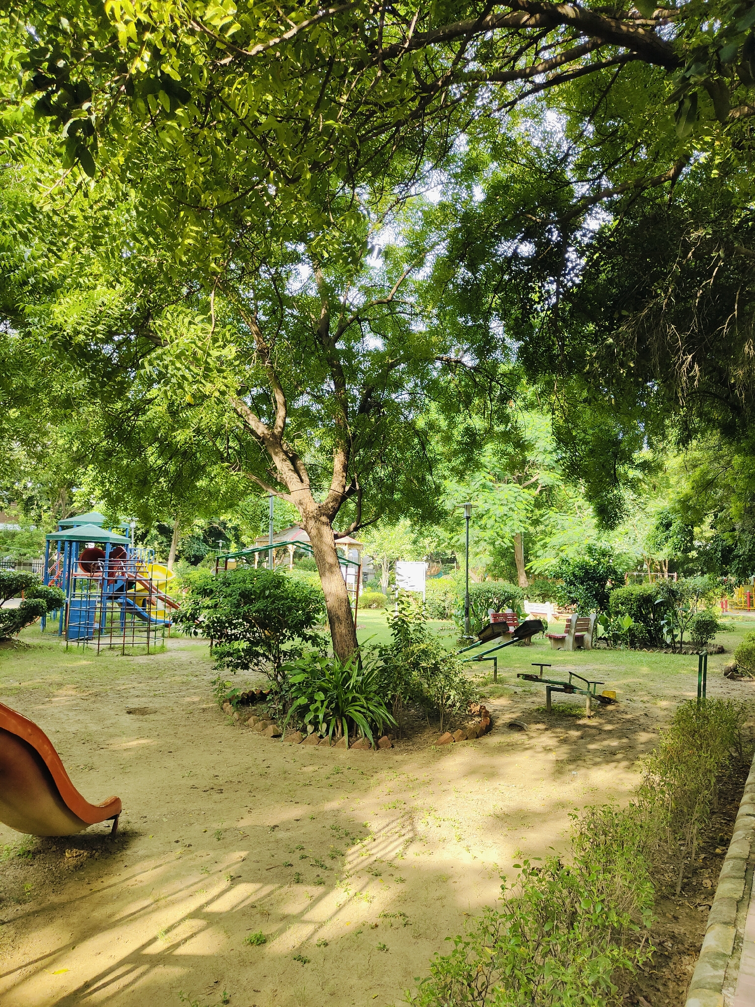 The image depicts a children's playground in a park in Gurgaon, India. The playground features a slide, swings, and climbing structures. The ground is covered with sand and dry leaves, and there are trees and bushes throughout the park. The park appears to be well-maintained, and the playground equipment is in good condition. The image was taken on a sunny day, and the lighting suggests it is either morning or afternoon. There are no people visible in the image, but the presence of playground equipment implies that it is a place for children to play.