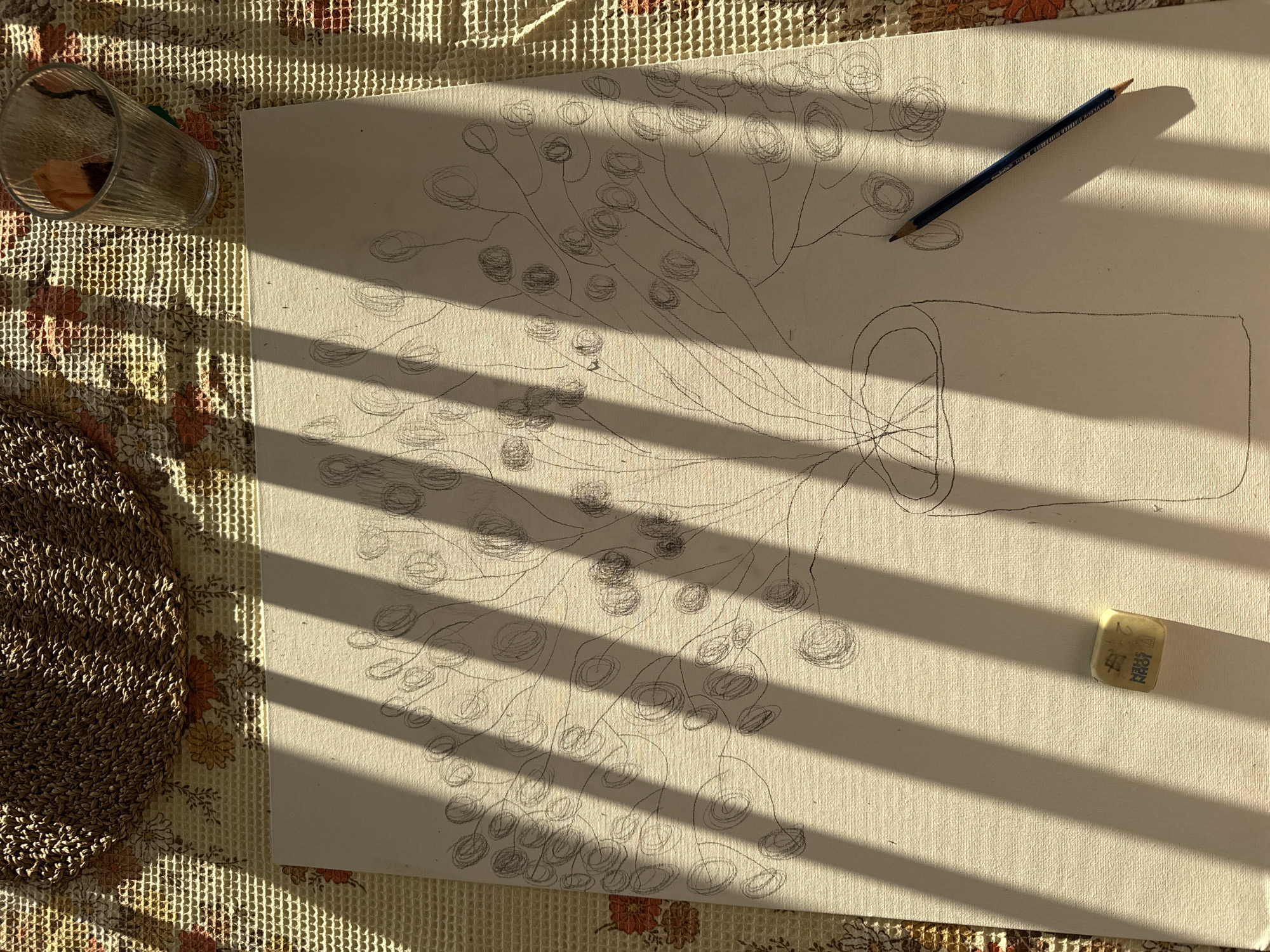 An overhead photograph taken in Kfar Vradim, Israel, captures an artistic workspace under strong, direct sunlight casting distinct, parallel vertical shadows across the scene. The central subject is a textured, off-white canvas featuring a graphite pencil sketch of a stylized plant arrangement in a cylindrical vase. The plant consists of multiple slender stems branching upwards, each terminating in a spiraled, circular form resembling a berry or bud. To the lower right of the canvas lies a blue pencil, while a white eraser with "JOHN SHEN" printed on it is positioned on the lower left. The canvas rests on a light-colored, patterned tablecloth with a floral design and a grid texture. In the upper left corner, a circular woven mat made of natural fibers is partially visible. A ribbed glass tumbler containing dried plant material is situated in the upper right quadrant of the image. No individuals are present.