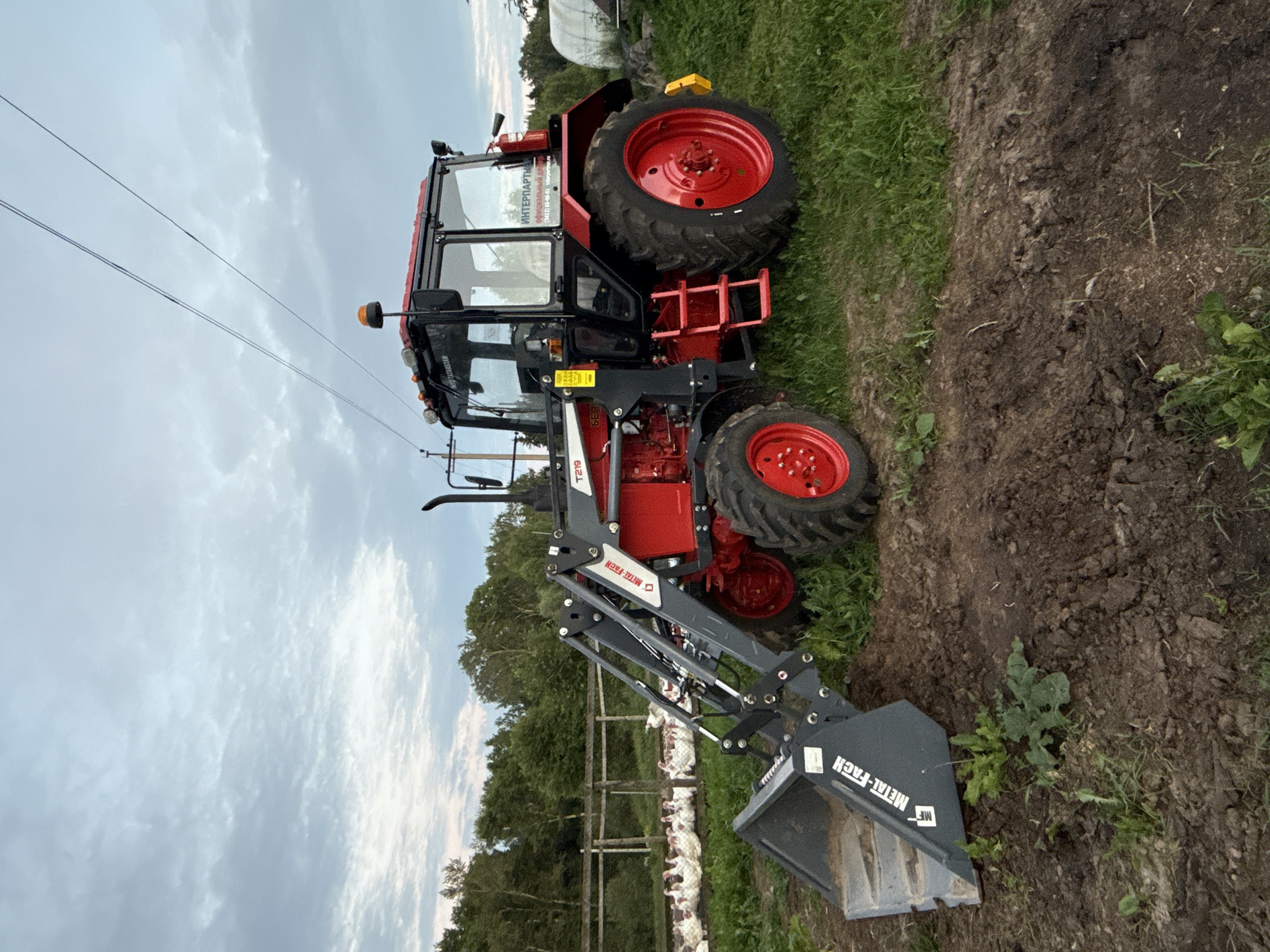 In the rural outskirts of Kukui, Russia, a vibrant red and black tractor with a front-end loader is parked on a patch of uneven, dark brown earth and green grass. The tractor features a fully enclosed cabin with tinted windows, an orange rotating beacon on its roof, and multiple exterior lights. The front loader is lowered, with its grey bucket resting on the ground, partially filled with soil and a small amount of green vegetation. The tractor's large rear wheels are bright red, contrasting with the black tires. In the background, a wooden fence encloses a flock of white turkeys resting on the grass. Beyond the fence, a dense line of green trees forms a natural backdrop. The sky above is overcast with soft, grey clouds, suggesting a late afternoon or evening setting. A single power line stretches across the sky above the tractor.