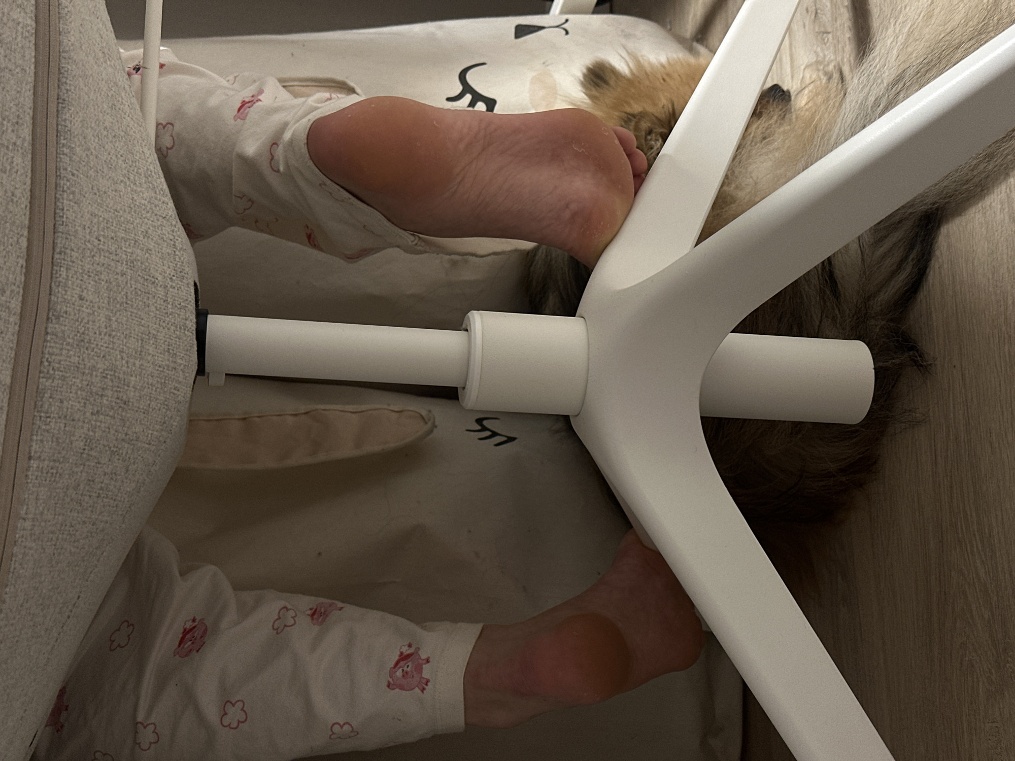 The image shows a person lying on a baby changing table. The person is wearing white pajamas with a pink pig and flower pattern. The feet of the person are visible. A white chair is positioned over the person's head. The changing table has a white surface with cartoon drawings. The setting appears to be indoors, possibly a bedroom. The exact location in Votkinsk, Russia is not discernible from the image.