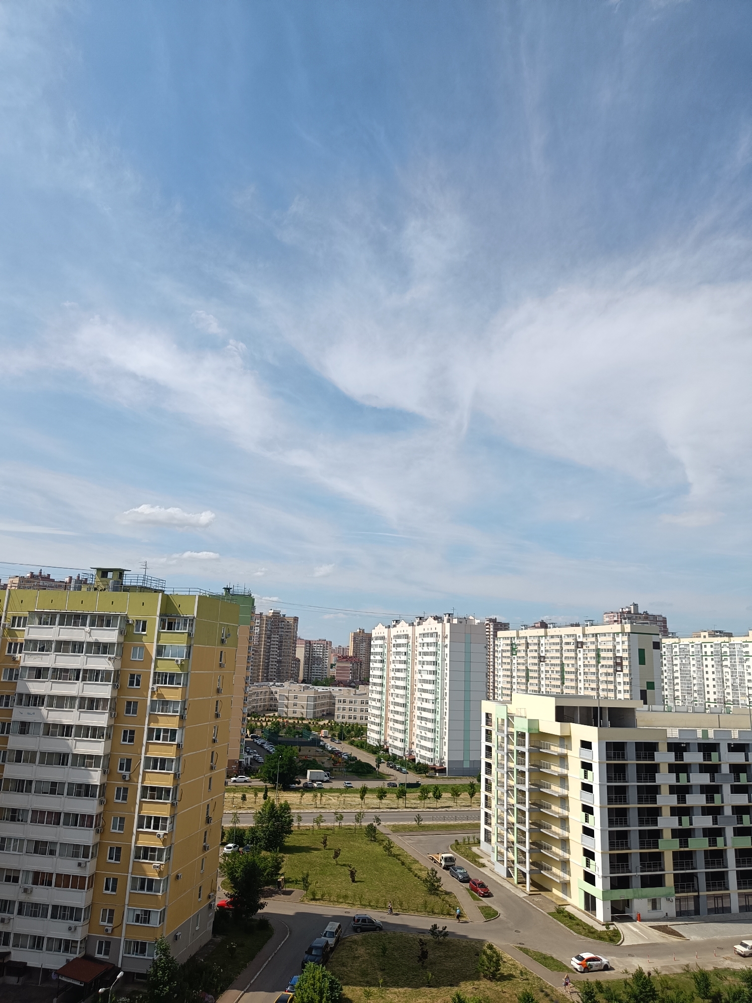 The image shows a panoramic view of a modern urban residential area on a bright, partly cloudy day. In the foreground, a tall, multi-story apartment building with a yellow and light green facade dominates the left side of the frame. Its exterior is characterized by numerous balconies and windows, indicating a high-density living environment.
To the right of the foreground building, a newer, under-construction or recently completed apartment complex is visible. This building features a more contemporary design with a light yellow, gray, and green color scheme, and exposed exterior staircases.
In the background, a dense cluster of high-rise apartment buildings stretches across the horizon, showcasing a variety of architectural styles and heights, typical of a large city. The buildings are mostly light-colored, with some featuring balconies and uniform window patterns.
The ground level includes paved roads, sidewalks, green lawns, and scattered trees. Several cars are parked on the streets and in parking lots, and a few pedestrians can be seen walking on the sidewalks. The sky above is a vibrant blue with wispy white clouds that create a dynamic pattern.
While the provided information suggests the location is Krasnodar, Russia, the image itself does not contain any explicit geographical markers or recognizable landmarks that would confirm this specific location. The scene depicts a typical modern urban landscape found in many developed cities.