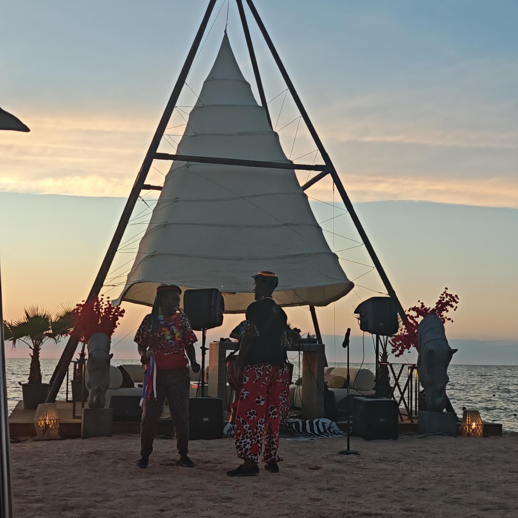 The image depicts an outdoor scene at what appears to be a beach or coastal area, with the ocean visible in the background under a twilight sky. In the foreground, two individuals are standing on sand, facing each other. The person on the left wears a red and black patterned shirt with a colorful lei and patterned pants. The person on the right is dressed in a black top, a red patterned headscarf, and red and white patterned pants, and is holding a microphone. Behind them is a large, triangular structure with white fabric resembling a sail, supported by a dark metal frame. Several speakers are positioned on either side of the stage area. To the left, a palm tree is visible. Decorative elements include what appear to be elephant statues and red floral arrangements. The scene suggests a performance or event is taking place. The location is identified as Мысовское сельское поселение, Ukraine.