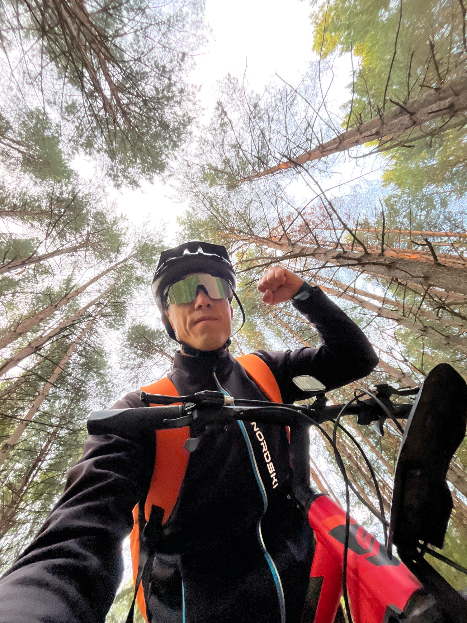 A low-angle, wide shot captures a male cyclist in a forest, looking up at the camera. The cyclist wears a black helmet, green cycling glasses, and a black jacket with an orange vest over it. He is holding the handlebars of a red and black bicycle and has his left fist clenched, possibly in a gesture of strength or triumph. The background consists of tall, bare trees with a bright sky visible through the branches. The environment suggests a natural, wooded area, likely outside of a populated urban center, such as a forest outside of Chaikovsky, Russia.