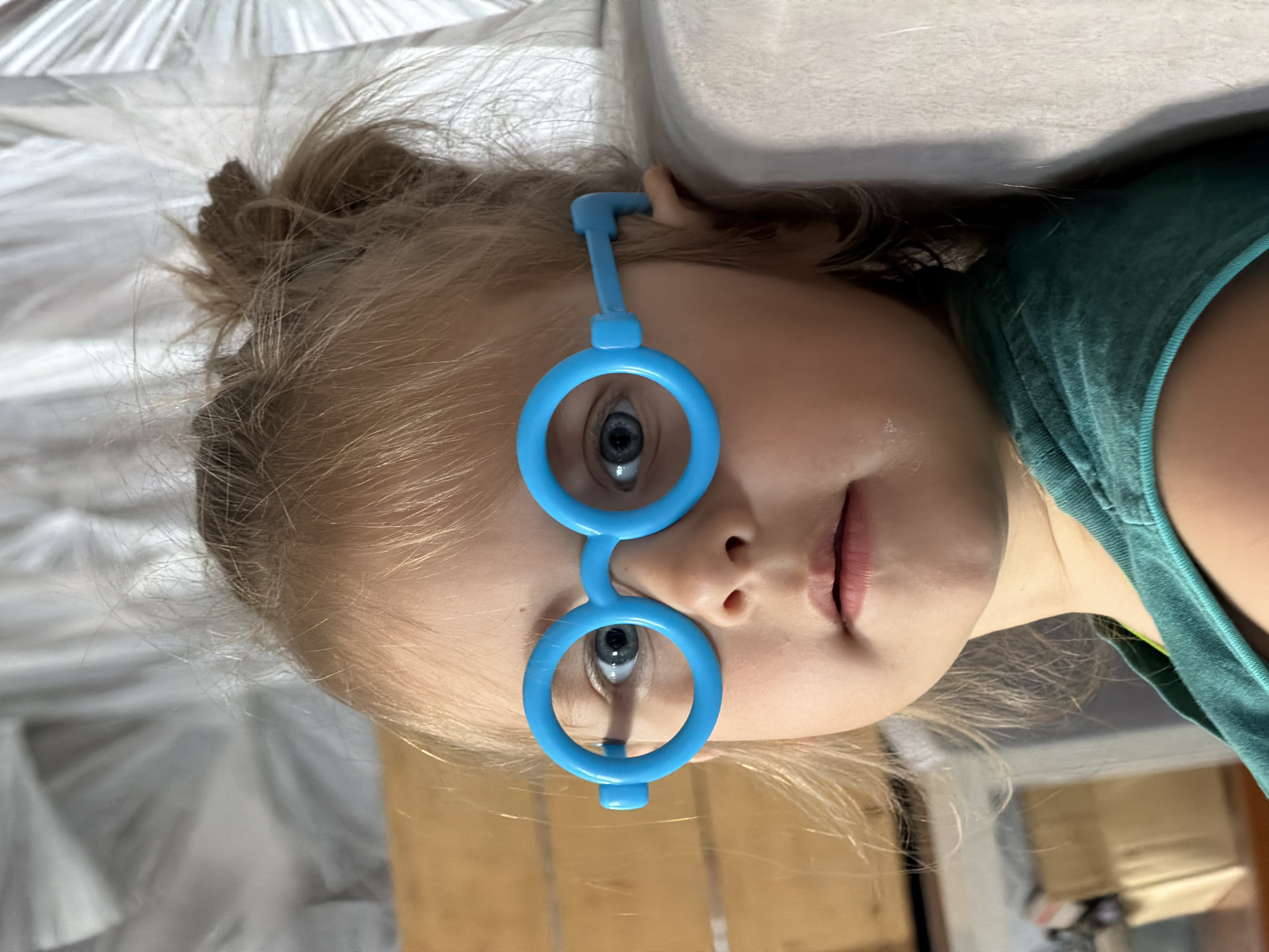 A close-up, eye-level shot shows a young Caucasian girl with blonde hair pulled into a messy bun, wearing large, bright blue toy glasses. She is looking directly at the camera with blue eyes and a neutral expression, her mouth slightly open. She is wearing a teal tank top. The background is out of focus, showing a textured white surface on the right and a wooden surface on the left. The lighting is natural and appears to be from the side. There is no indication of the city of Chaikovsky, Russia in the image.
