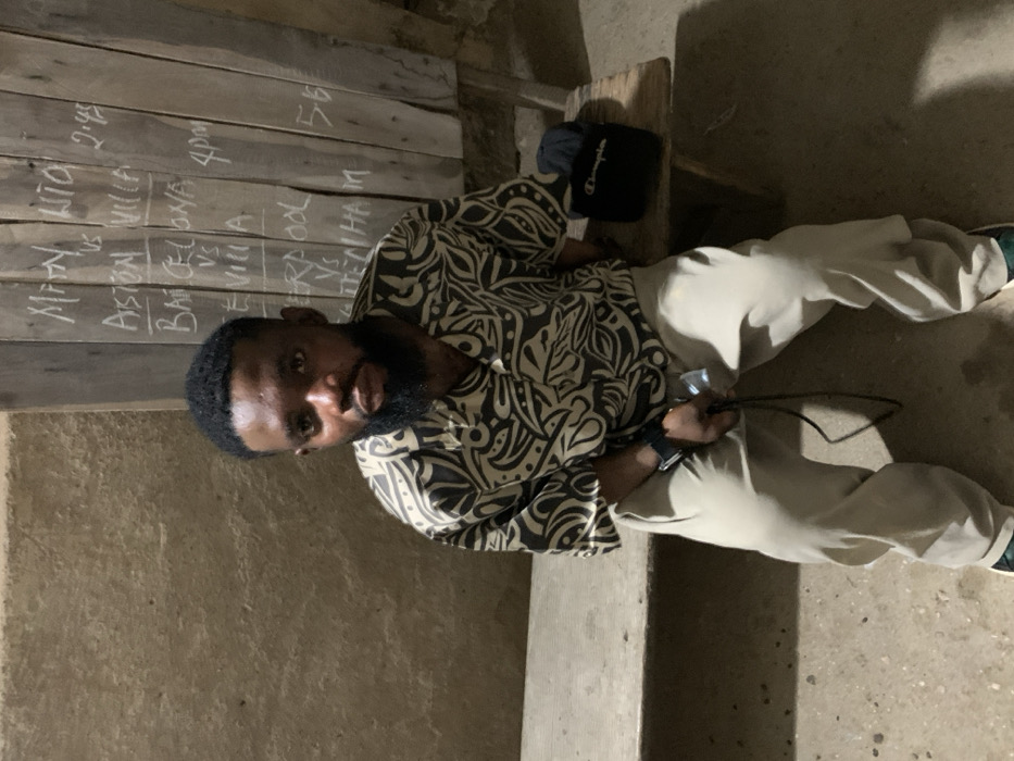 A dark-skinned male individual is seated on a rustic wooden bench in Okene, Nigeria. He wears a short-sleeved collared shirt with a black and light-patterned design, light-colored trousers, and a dark smartwatch on his left wrist. In his left hand, he holds a black cable with a silver metallic end. His gaze is directed forward. To his right, on the bench, rests a black pouch with "Champion" inscribed in white, next to a folded dark fabric item. The background features a textured, light brownish-grey concrete or mud wall on the left. On the right, a vertically planked wooden board displays handwritten chalk text listing football matches and times: "MAN UTD vs ASTON VILLA 2:45", "BARCELONA vs SEVILLA 4pm", and "LIVERPOOL vs TOTTENHAM 5:6". The scene is dimly lit, suggesting an indoor or covered setting.