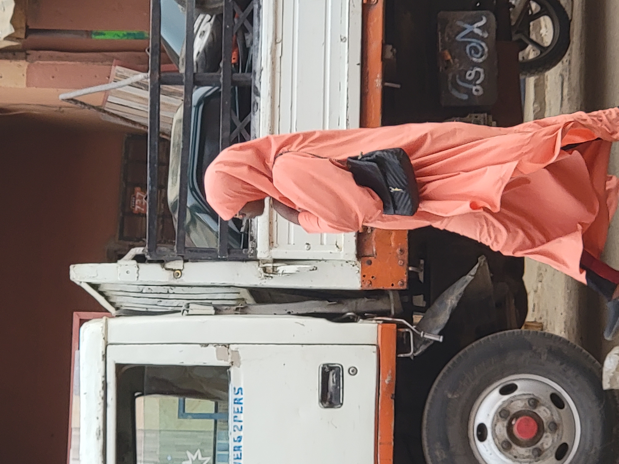This image captures a street scene in Maiburiji, Nigeria, under bright daylight. The primary subject is a person dressed in a flowing, peach or light orange-colored garment and matching head covering, with a black shoulder bag slung across their chest. Their head is bowed, and their face is largely obscured as they stand beside a truck.
The truck, positioned prominently in the frame, features a white cabin and a functional, utilitarian design with orange accents on its chassis or cargo bed. It appears to be a well-used vehicle, possibly a small utility truck or pickup. On the side of the truck's cabin, blue text is visible, reading "VERS 2PERS," likely indicating "2 PERSONS" or "2 PASSENGERS."
In the background, behind the truck and the person, hints of other structures and activities are visible. A dark, rectangular sign or surface in the upper right background bears white, chalk-like graffiti that appears to spell "Xo So" or similar stylized characters, next to which a motorcycle wheel and tire are partially seen. The overall setting suggests a bustling outdoor environment, possibly a street or market area, with muted earthy tones dominating the background elements.