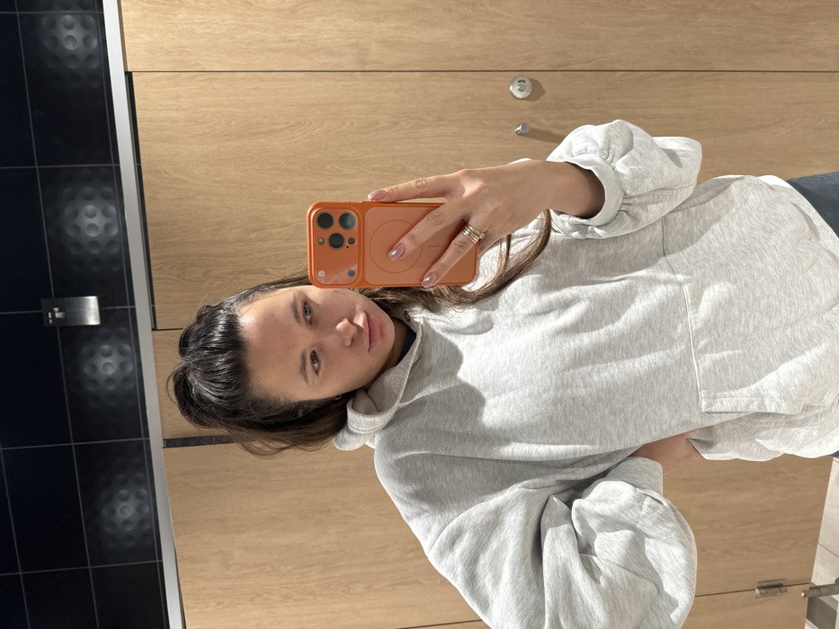 A young woman poses for a selfie in a public restroom. She holds an orange smartphone with her right hand, which has multiple rings on the ring finger. She wears a light grey hoodie with balloon sleeves and a pocket. Dark hair is partially pulled back. The background shows a wall of light woodgrain paneling and a section of dark, textured tile.