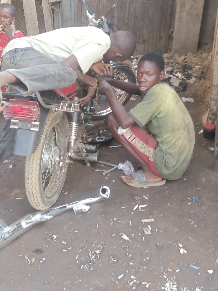 Two young men work on a motorcycle. One leans over the bike, appearing to work on the engine. The other squats nearby, looking at the camera. A new chrome exhaust pipe lies on the ground next to various tools and debris. The scene appears to be an outdoor motorcycle repair shop, with a child visible in the background.