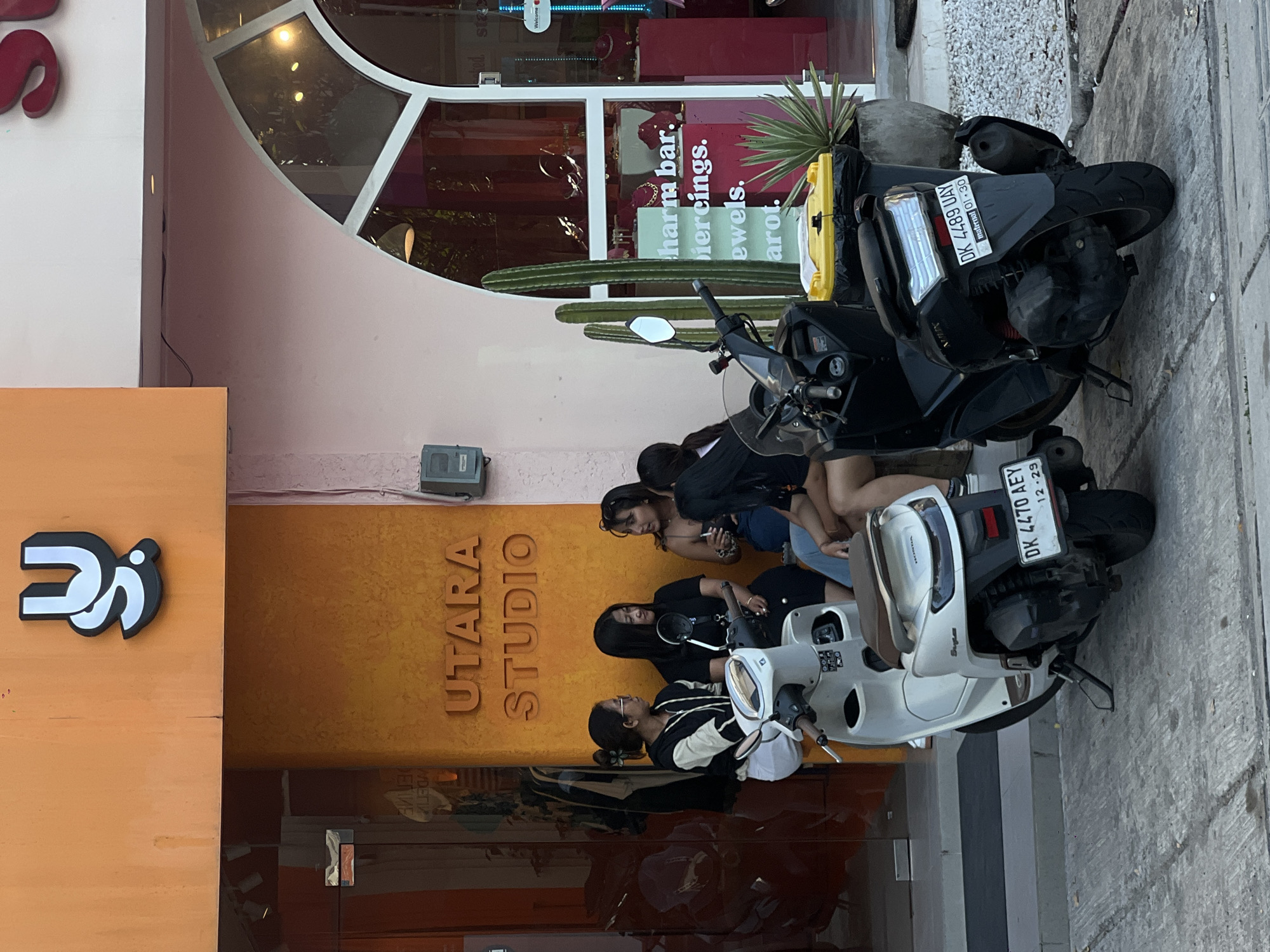 The image depicts the exterior of a business named "UTARA STUDIO" located in North Kuta, Indonesia. The studio has a storefront with a large arched window above a sign that reads "charm bar, piercings, jewels, tarot." In front of the building, there are several parked scooters. Three young women are gathered around one of the scooters. Two scooters are visible with license plates: DK 4470 AEY and DK 4489 UAY. Cacti decorate the entrance.