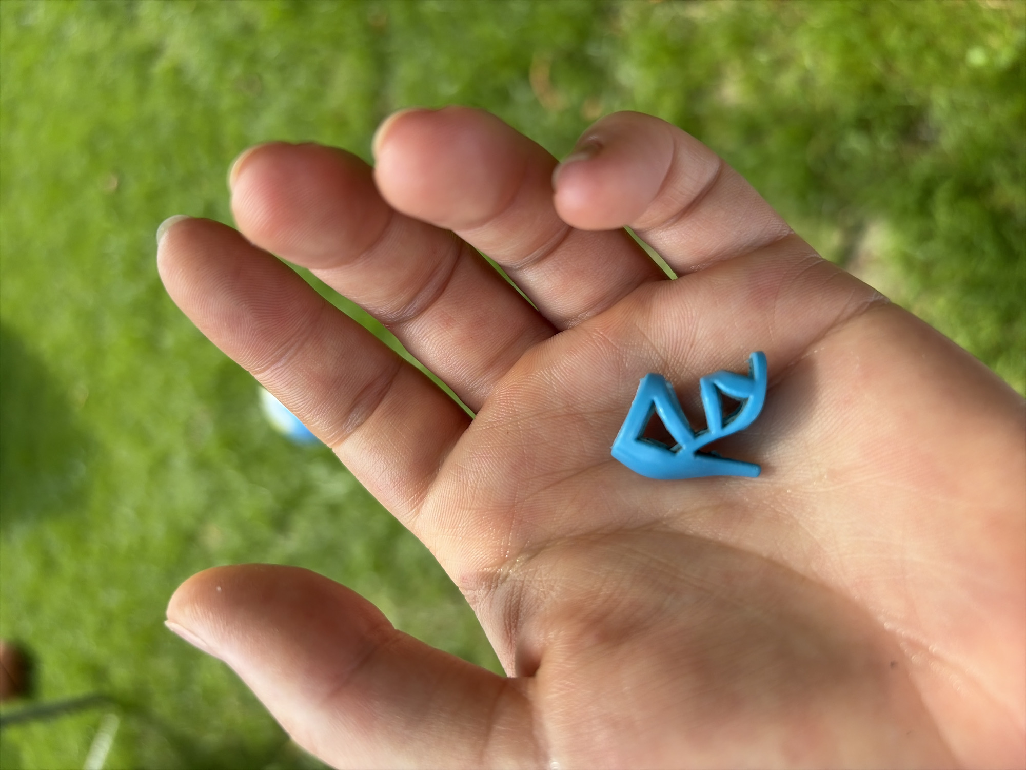 A hand is holding a small blue plastic high-heeled shoe ornament. The hand is open with fingers slightly curled. The ornament is positioned on the palm. The background is blurred green grass. The scene is outdoors on a sunny day. There is no indication of the location being Oxentea, Moldova.
