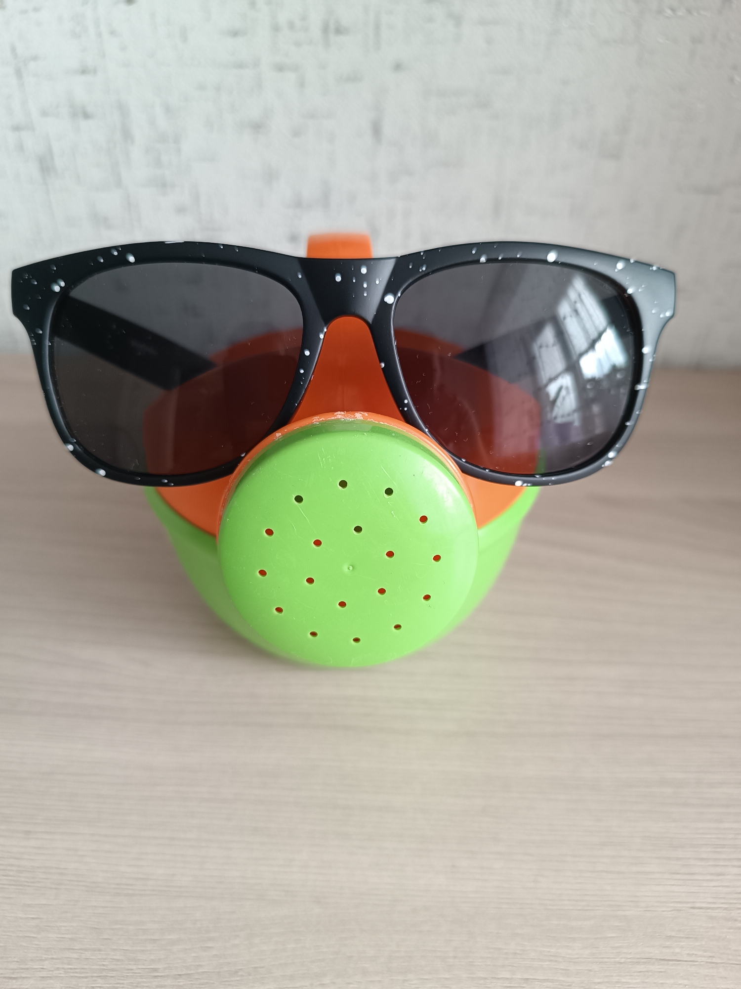 A still-life image shows a green and orange watering can indoors with black sunglasses placed on top, creating a face-like appearance. The watering can is resting on a light-colored surface, possibly a table or countertop. The background is a textured wall. The location is Chaikovsky, Russia.
