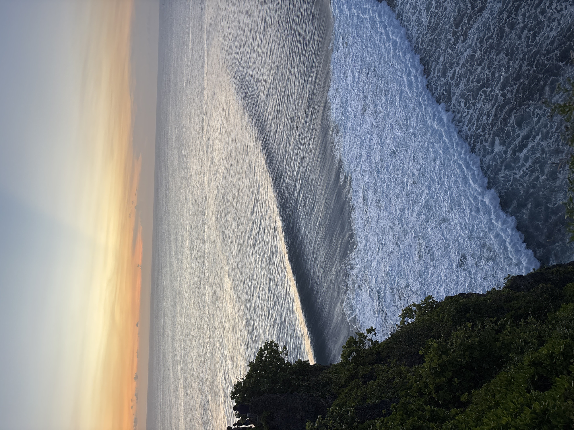 The image depicts a view from a cliff overlooking the ocean in Kuta Selatan, Indonesia. A wave is breaking along the coastline, with foamy white water contrasting with the calmer, silvery surface of the sea. The sky transitions from a warm, yellowish-orange hue on the left to a pale blue-gray further out. Dense, green vegetation covers the edge of the cliff in the foreground. Two figures are visible on the wave.