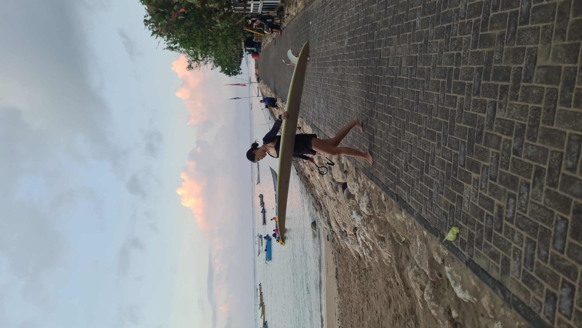 In Denpasar, Indonesia, a woman walks barefoot along a stone-paved promenade adjacent to a sandy beach. She is carrying a long surfboard horizontally. The ocean is to her left, with several small boats anchored in the water. The sky above is a pale blue with scattered, wispy clouds tinged with pink from what appears to be sunrise or sunset. Vegetation and structures are visible on the right side of the promenade.