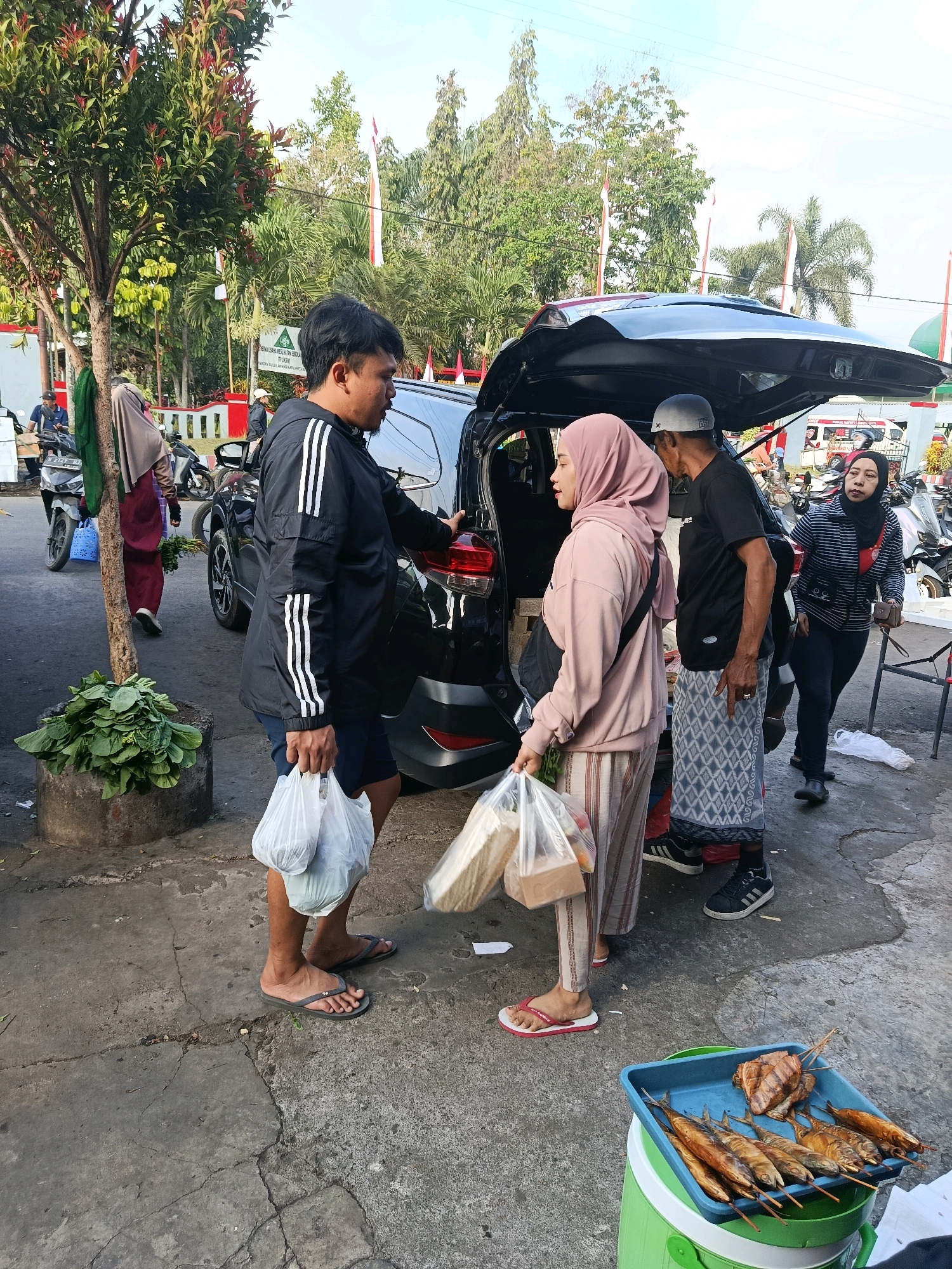 A man in a black Adidas jacket and blue shorts stands facing a woman wearing a pink hijab and a long-sleeved pink hoodie with striped pants. He holds two plastic bags of groceries. The woman holds two plastic bags. Behind them, a black SUV's trunk is open. To the right of the SUV, a man in a black shirt and a patterned sarong stands, wearing a grey skullcap. Further to the right, a woman in a striped hijab and top is visible. In the foreground, a blue tray filled with grilled fish is placed on a green cooler. In the background, a tree with red-tipped leaves is on the left, and several motorcycles are parked. Red and white flags are visible in the distance. The scene appears to be an outdoor market in Bululawang, Indonesia, with a group of people interacting near a vehicle, likely engaged in shopping.