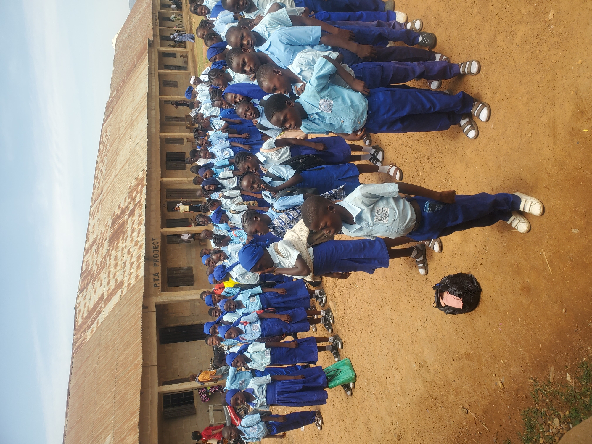Akwanga, Nigeria: A large group of African school children, dressed in blue uniforms and sandals or shoes, are standing in rows on a red dirt ground. The children are facing forward, with their hands clasped in front of them. In the background is a building with a corrugated metal roof and the words "P.T.A. PROJECT" written on the side. A black backpack is on the ground nearby.