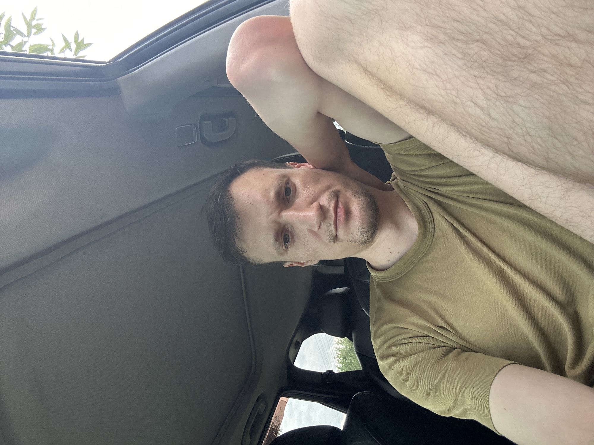 A young man with short brown hair and light skin is sitting in the driver's seat of a car, looking directly at the camera. He is wearing a light brown t-shirt. His left arm is bent and resting on his knee. The interior of the car is visible, including the headliner, a sunroof, and the seats. Outside the car, trees and a cloudy sky can be seen through the windows. The image appears to have been taken in the city of Makiv, Ukraine.