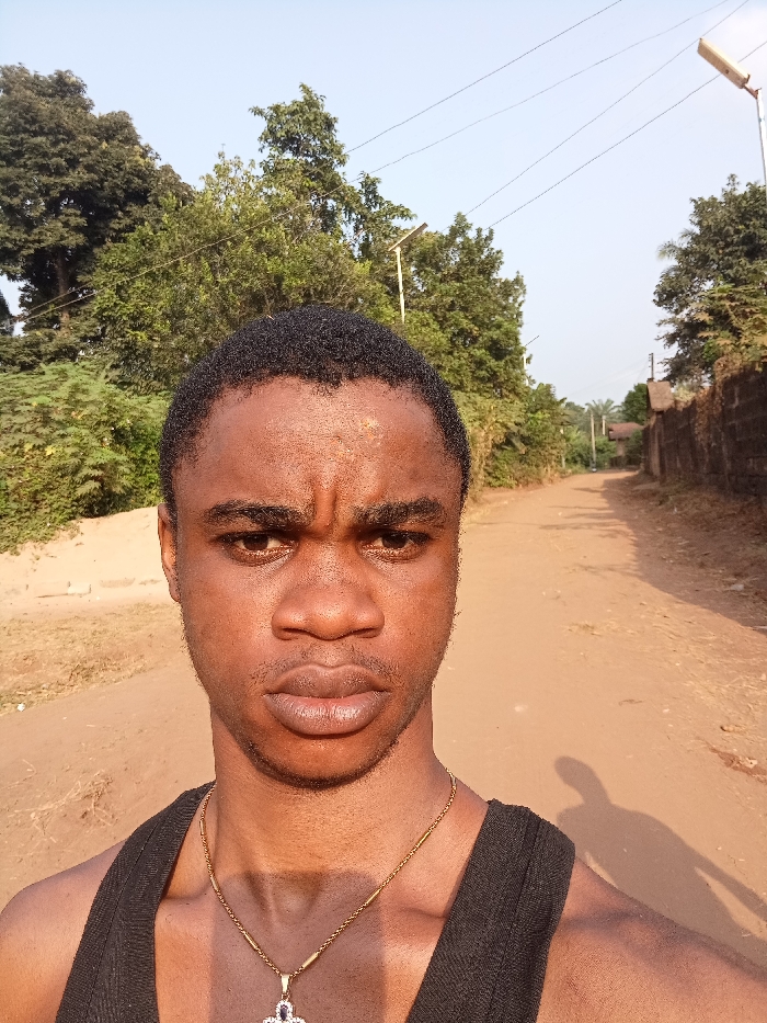 A young man takes a selfie on a dirt road. He is wearing a black tank top and a gold necklace with a cross pendant. He has a serious expression. The road stretches behind him, flanked by trees and a wall. Utility poles line the road. The sunlight creates a shadow of the man's arm and phone.