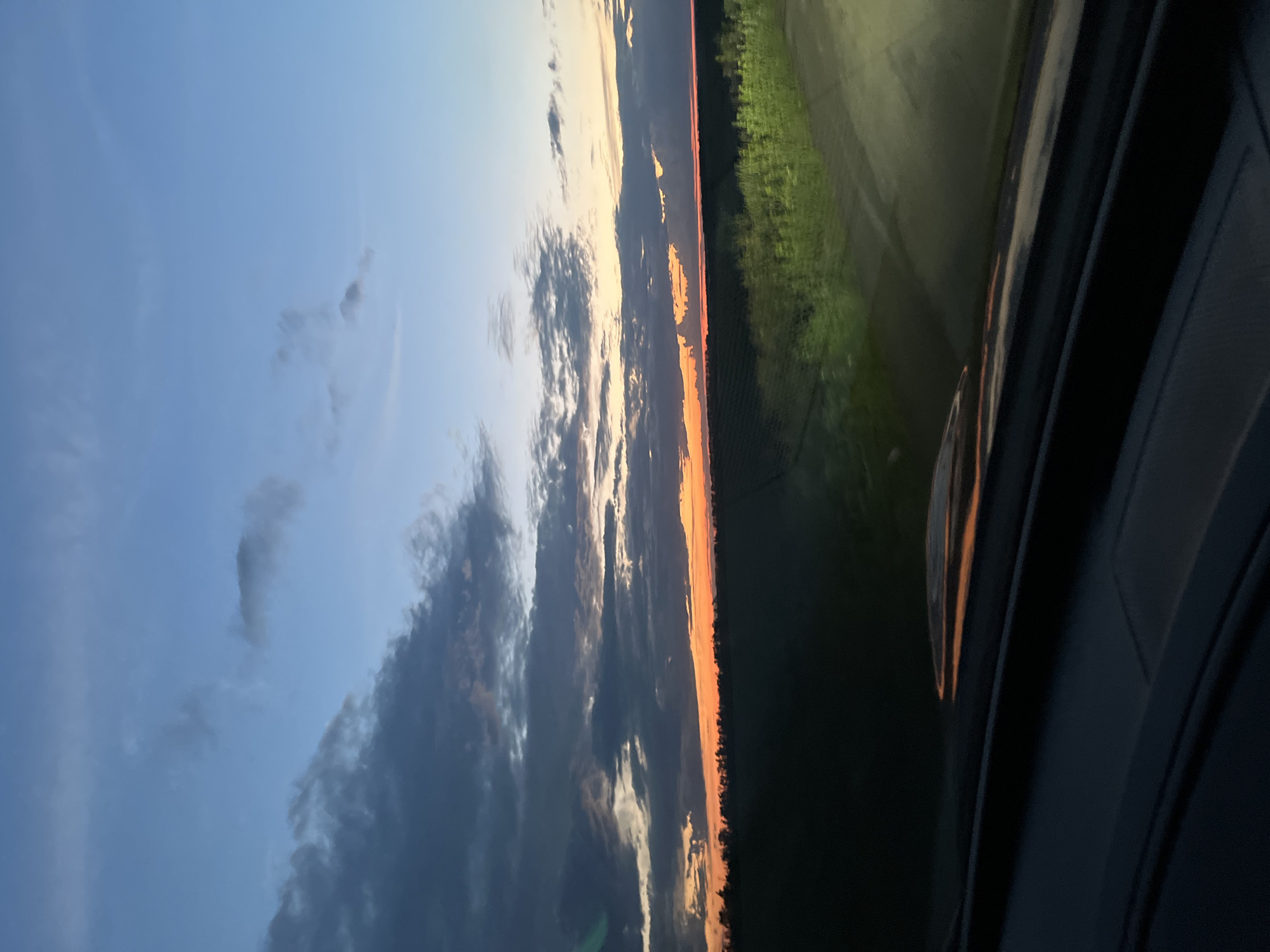 The image depicts a rural landscape at dusk, likely captured from a vehicle. The sky is a gradient of blues and oranges, with scattered clouds. The horizon is lined with a dark silhouette of trees against the vibrant sunset. In the foreground, the interior of a car is visible, including the dashboard and a windshield reflecting the twilight sky and surrounding greenery. The fields are dark, with some areas illuminated by what appears to be headlights or artificial lighting, casting a green glow on the vegetation. The location is identified as Poltavka, Ukraine, suggesting an agricultural or undeveloped area. There are no people or animals visible in the scene. The overall mood is tranquil and serene, characteristic of a sunset in a rural setting.