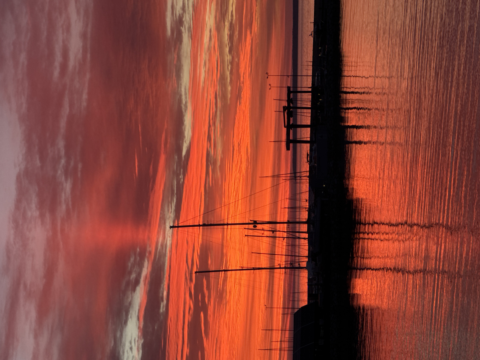 A vibrant sunset paints the sky over the Baltic Sea in Tallinn, Estonia, casting a fiery glow on a marina filled with silhouetted sailboats. The clouds are a dramatic tapestry of oranges, reds, and pinks, with a distinct ray of light piercing through the center. The calm water reflects the vivid colors of the sky and the masts of the sailboats, creating a mirror image of the scene above. In the foreground, the marina is dark and indistinct, with the outlines of boats and structures visible against the bright sky. The overall impression is one of serene beauty and natural splendor.