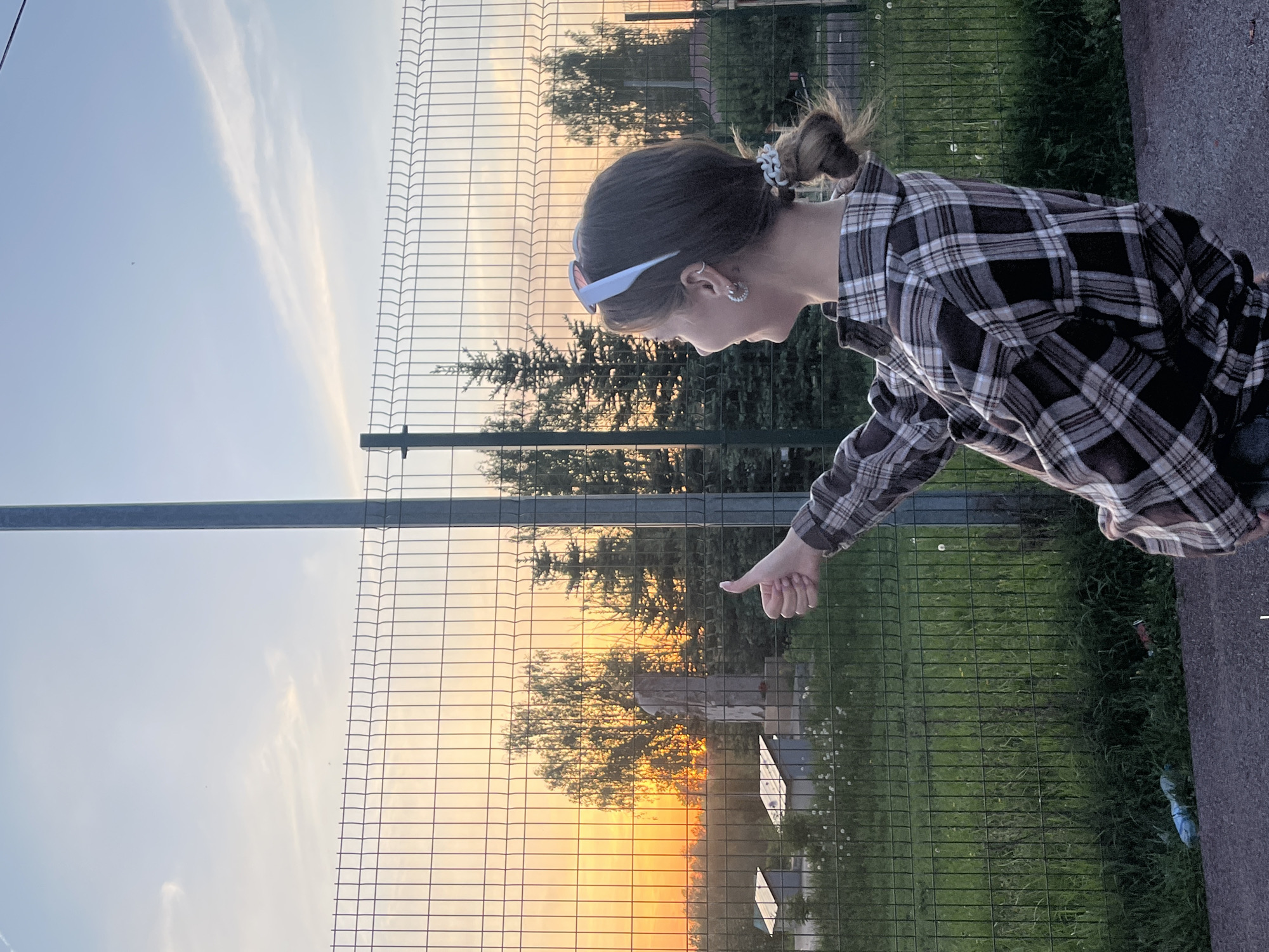 A person with a bun hairstyle and wearing a plaid shirt is standing next to a green wire fence. The person is holding their thumb up. Behind the fence, there is a sunset and some trees. The location is Фоки, Russia.