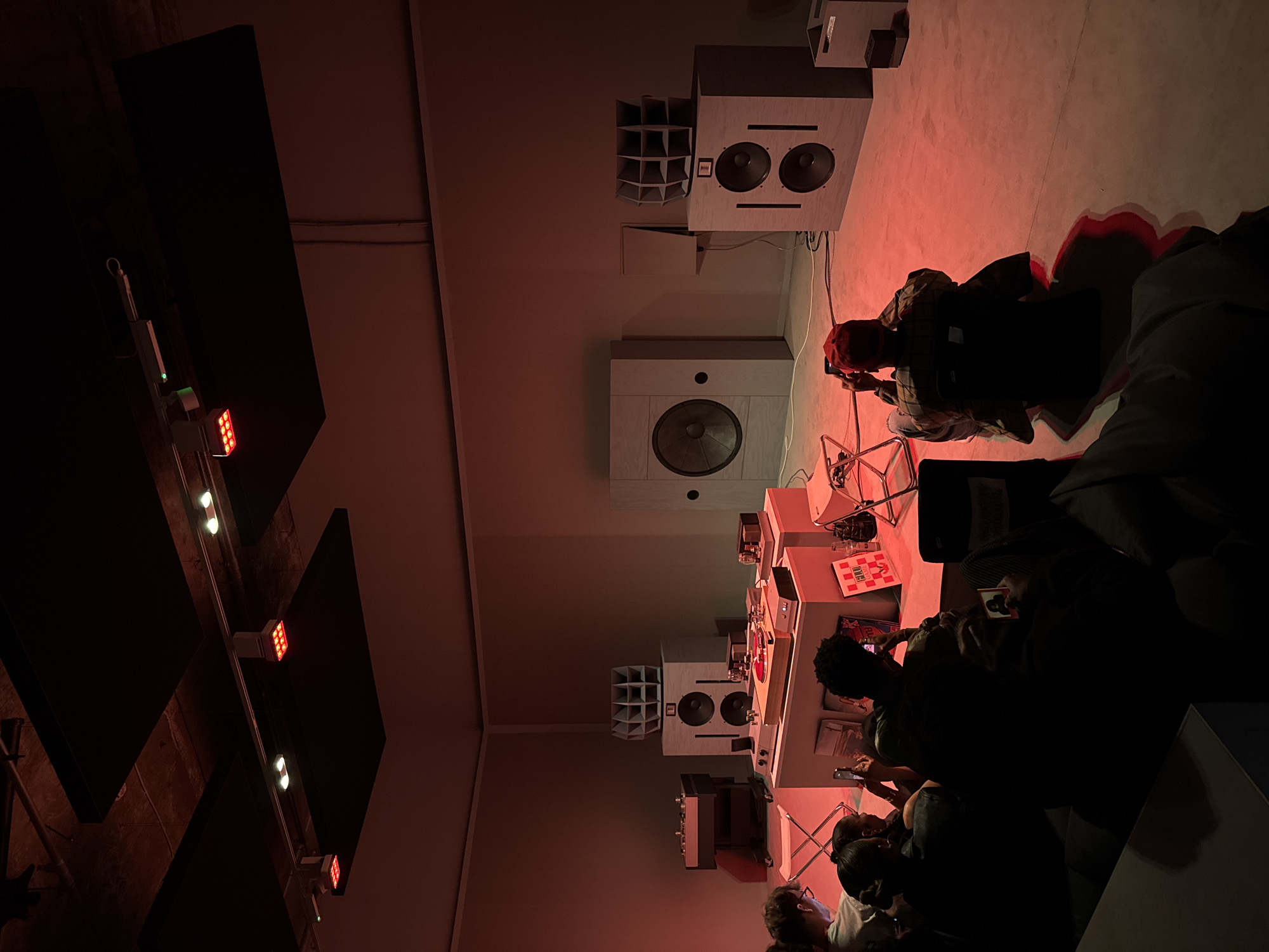 The image depicts an indoor space, likely a venue or studio in London, United Kingdom, characterized by a reddish hue due to the lighting. The space is fitted with a series of large, rectangular loudspeakers mounted on the walls, including models with single and dual woofers. Several people are seated on the floor and on chairs, focused on the tables in front of them, possibly engaged in activities involving the equipment there. The tables feature vinyl records, mixers, and other electronic devices, suggesting a DJ set or music production scenario. Above, the ceiling has dark acoustic panels with strategically placed lights. The overall atmosphere is that of a creative event or workshop with a focus on music.