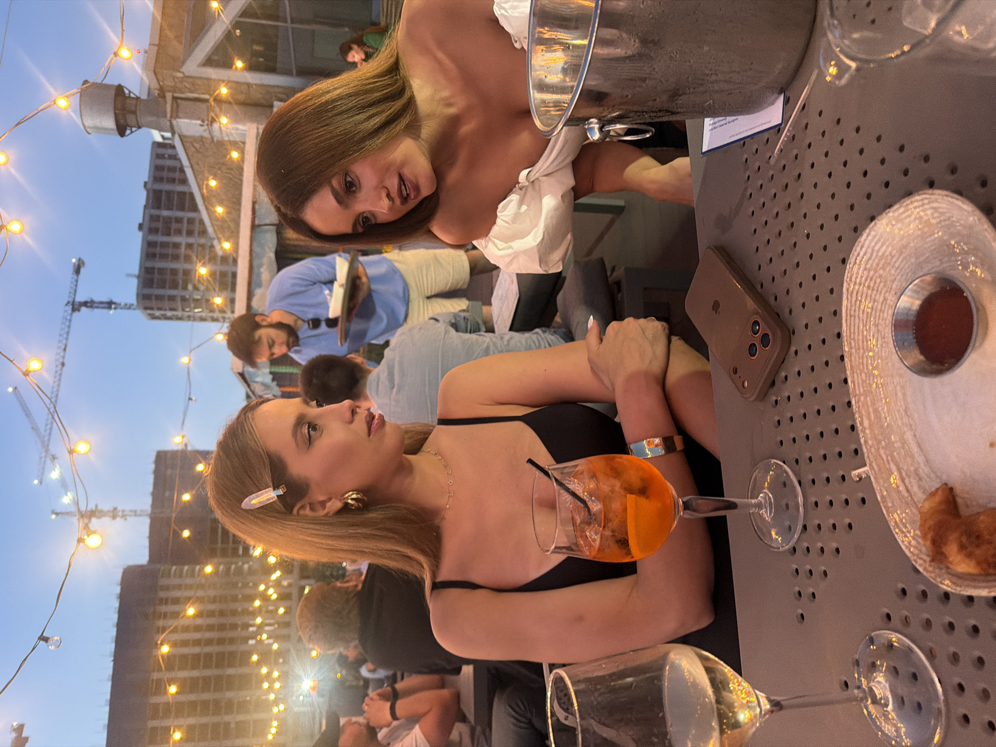 This media file depicts a group of people gathered on a rooftop setting, likely for a social event. The scene is illuminated by string lights, suggesting it is evening or dusk. In the foreground, a woman with blonde hair is looking to the left, wearing a black top and a bracelet. She has a drink with orange liquid and ice in a stemmed glass. Another woman, with brown hair, is visible in the background, leaning forward and looking towards the viewer.
The background shows urban elements, including tall buildings and a construction crane, indicating the location is a city. The overall atmosphere appears to be relaxed and social, with people casually dressed and engaged in conversation. The presence of drinks and what looks like food items on the table suggests a meal or a gathering at a restaurant or bar. The weather is not explicitly discernible but the lighting suggests it's not a bright sunny day. There is no visible text in the image.