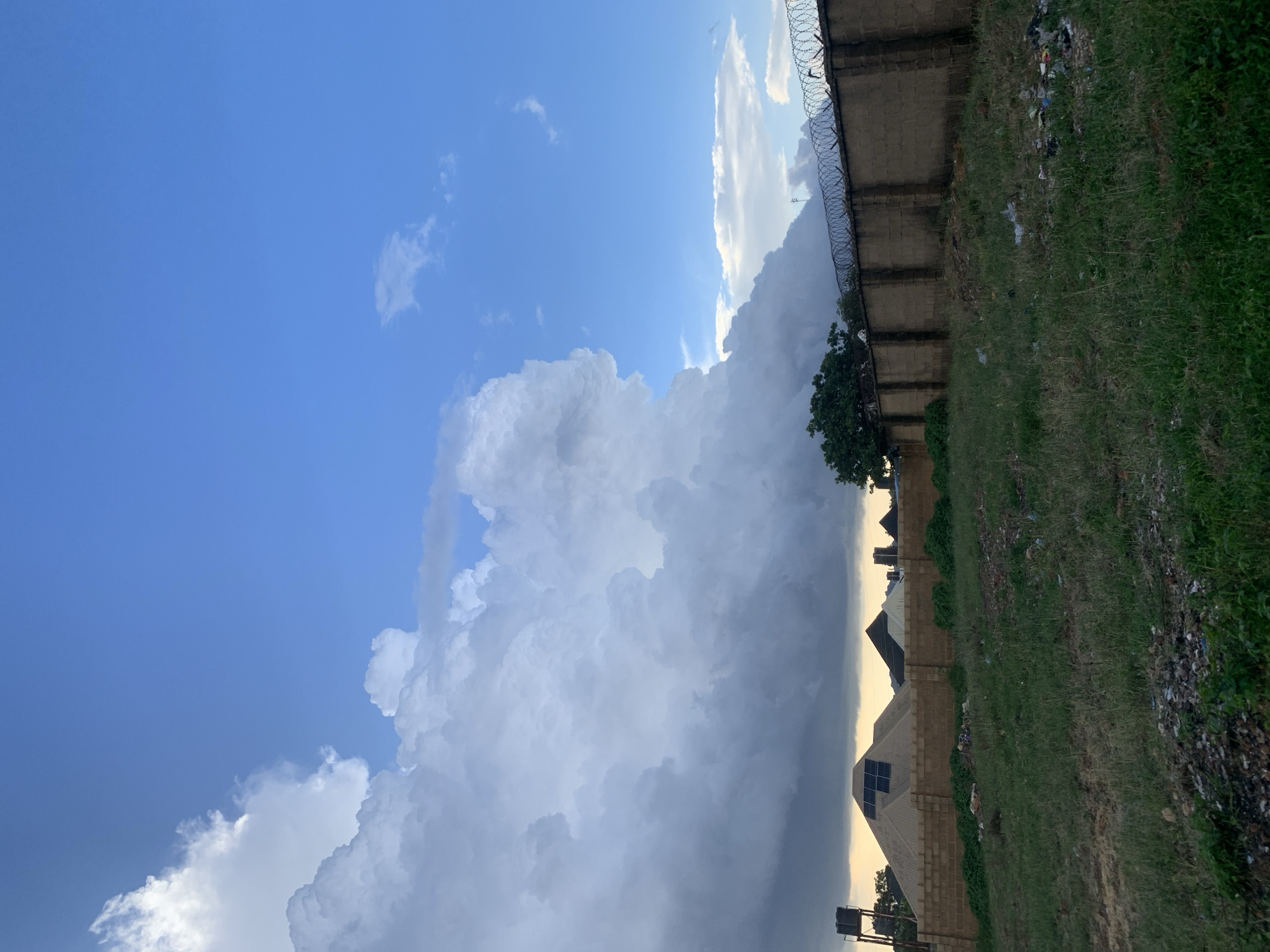 The image depicts an outdoor scene in Rafin Bauna, Nigeria, with a focus on the sky and the immediate surroundings. The sky is a dominant feature, with a bright blue expanse on the left transitioning into a dramatic display of large, white, and grey clouds that occupy the center and right of the frame. The clouds suggest impending weather, possibly rain or a storm. The lighting indicates it is either early morning or late afternoon, with the sun casting a warm glow behind the clouds on the horizon.
In the foreground and along the right edge of the image, a grassy area is visible, interspersed with some scattered debris and a concrete wall topped with barbed wire, suggesting a boundary or enclosure. Behind the wall, the rooftops of several buildings are visible, some with what appear to be solar panels. A single tree with green foliage stands near the wall. The scene is still and quiet, with no people visible. The overall impression is one of a natural landscape under a dynamic sky, with human elements present but not the primary focus.