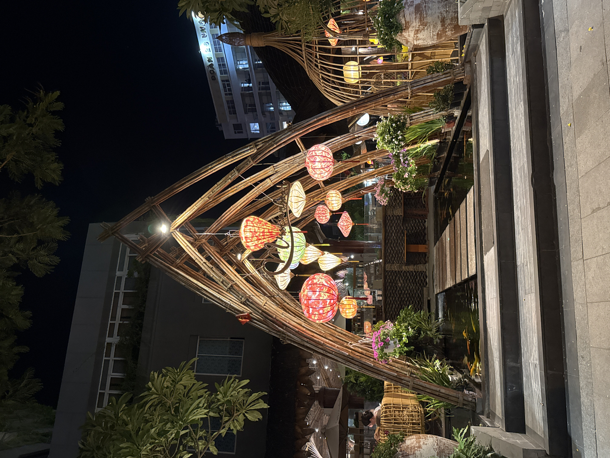 This image captures an outdoor scene at night, likely in Nha Trang, Vietnam. The primary focus is a decorative bamboo structure adorned with numerous colorful lanterns, creating a vibrant and festive atmosphere. These lanterns, in shades of red, orange, green, and white, are illuminated, casting a warm glow. The bamboo structure itself is intricately designed, resembling an arched entryway or canopy, with smaller bamboo poles forming an elaborate framework.
Behind the bamboo structure, a multi-story building with many windows is visible, suggesting an urban or semi-urban setting. Trees with lush green foliage are scattered throughout the scene, adding a natural element to the otherwise man-made environment. In the foreground, stone steps and a dark, reflective surface, possibly a pool or water feature, are present. The water seems to reflect some of the light from the lanterns.
There are a few people visible in the background, appearing to be patrons or staff of an establishment. One person is partially visible sitting near a wicker basket. The overall impression is one of a lively evening ambiance, possibly at a restaurant, cafe, or cultural event. The time of day is clearly night, indicated by the dark sky and the prominent illumination of the lanterns. The weather appears to be clear.
Visible text on the building in the background reads "DOAN 23 NHA".