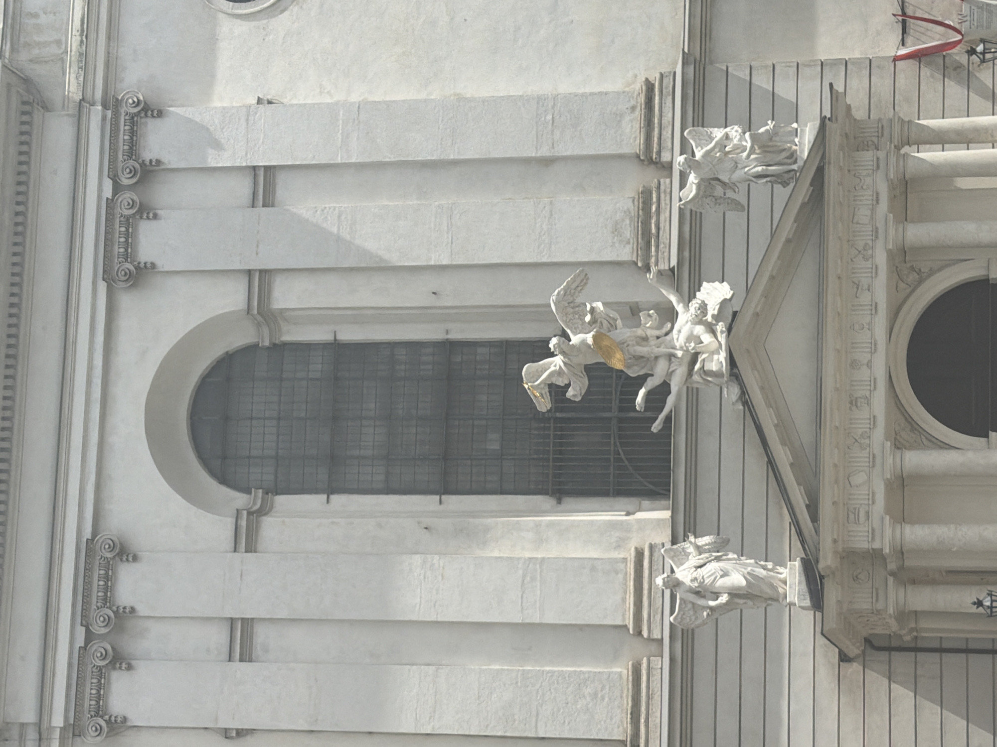 This is a medium shot, vertically oriented, exterior photograph of the facade of a grand building, likely a church or historical edifice, in Vienna, Austria. The building is constructed of light-colored stone, possibly marble or limestone, and features classical architectural elements. The facade is characterized by horizontal bands of stone, pilasters with ornate Ionic capitals, and a large, arched window with a metal grate.
The primary subjects are several white stone sculptures depicting angelic figures. In the upper portion of the image, an angel with outstretched wings appears to be in motion, possibly ascending or descending. Below this, a more complex sculpture features multiple angelic figures engaged in what appears to be a narrative scene. One angel, with golden elements, is particularly prominent. In the lower right of the image, another angelic sculpture is visible, appearing to be a grotesque or gargoyle-like figure. These sculptures are intricately detailed, showcasing flowing drapery and expressive poses.
The scene is set outdoors, under what appears to be bright sunlight, casting distinct shadows across the textured stone surfaces. The time of day is likely midday or early afternoon, given the intensity of the light and the shadows. The weather appears clear and sunny.
Notable details include the architectural style, suggesting a Baroque or Neoclassical period. The use of marble or a similar stone indicates a high-quality construction. The presence of the golden element on one of the sculptures adds a touch of richness. The building's facade suggests it is a significant historical landmark.
There is no visible text in the image. The overall emotion conveyed by the image is one of grandeur, artistry, and perhaps solemnity due to the religious nature of the sculptures.
