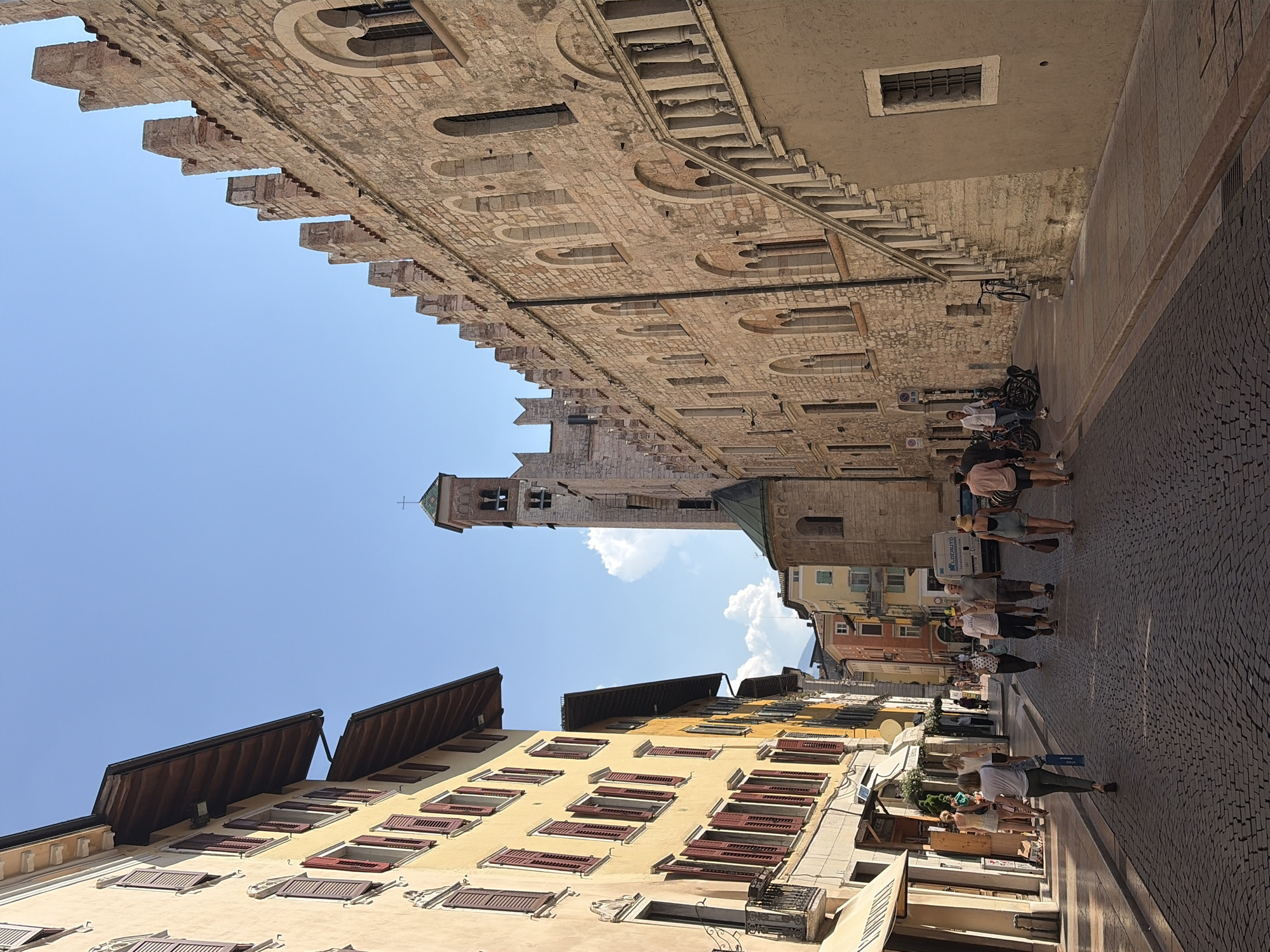 The image depicts a street scene in Trento, Italy. On the left, a yellow building with multiple windows and red shutters lines the street. To the right, a large, old stone building with arched windows and crenellations dominates the view, featuring a prominent stone staircase. In the background, a tall bell tower with a green roof and a cross can be seen. Several people are walking along the cobblestone street, some individually and others in small groups. One person is carrying a blue bag, and a few individuals are carrying shopping bags. Bicycles are parked near the stone building. The sky is clear and blue, with some scattered white clouds. The environment appears to be a historic city center with well-maintained architecture.