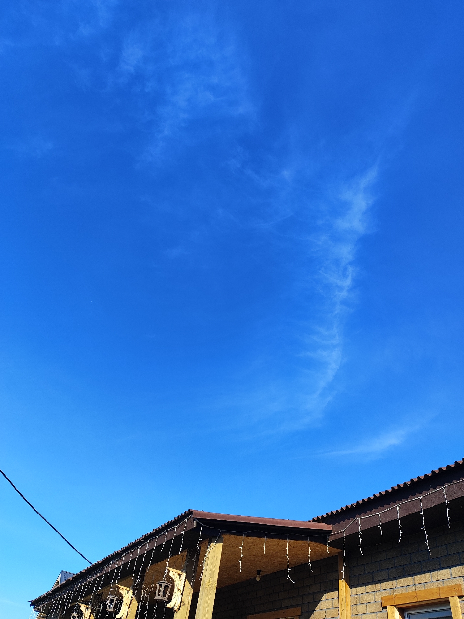 The image shows a clear blue sky with a wispy white cloud formation extending vertically from the top right towards the center. Below the sky, the edge of a building's roofline is visible, extending from the left side of the frame towards the right. The roof is covered with dark corrugated material and adorned with a string of white icicle-shaped lights. Some wooden support beams are visible under the eaves. The wall of the building is constructed with brown brickwork and some wooden trim. A black electrical wire is strung diagonally across the upper left portion of the sky. The setting appears to be outdoors, possibly in a residential area in Marusino, Russia, given the context. No people are visible in the image.