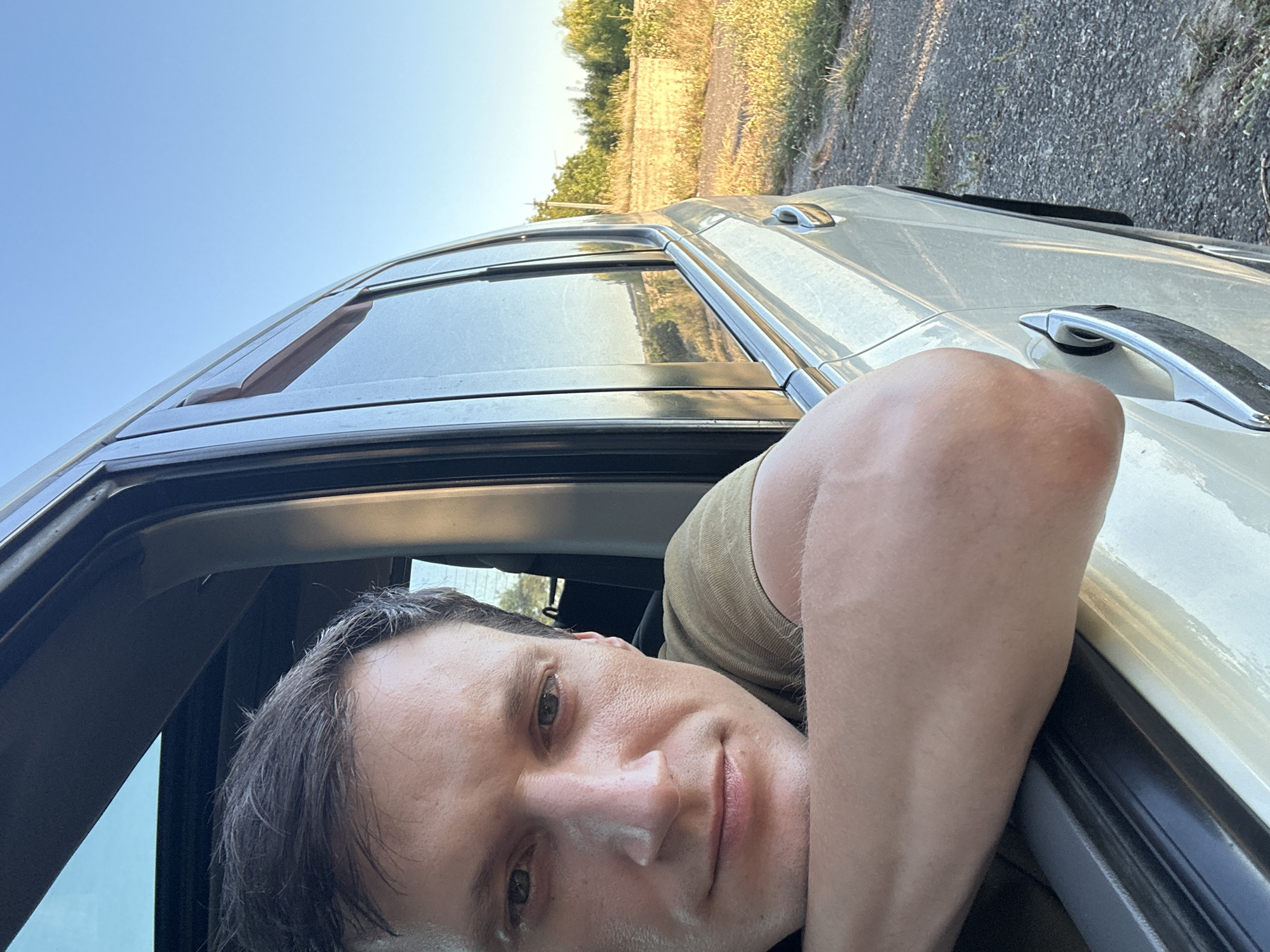 The image shows a man leaning out of a car window, resting his head on his arms. The car is light-colored and appears to be stationary. The man has short, dark hair and is wearing a light-colored t-shirt. He is looking directly at the camera with a neutral expression. The background is a rural landscape with a field of dry grass and trees under a clear blue sky. The sun appears to be low in the sky, casting long shadows. The location is identified as Makiv, Ukraine, based on contextual information.