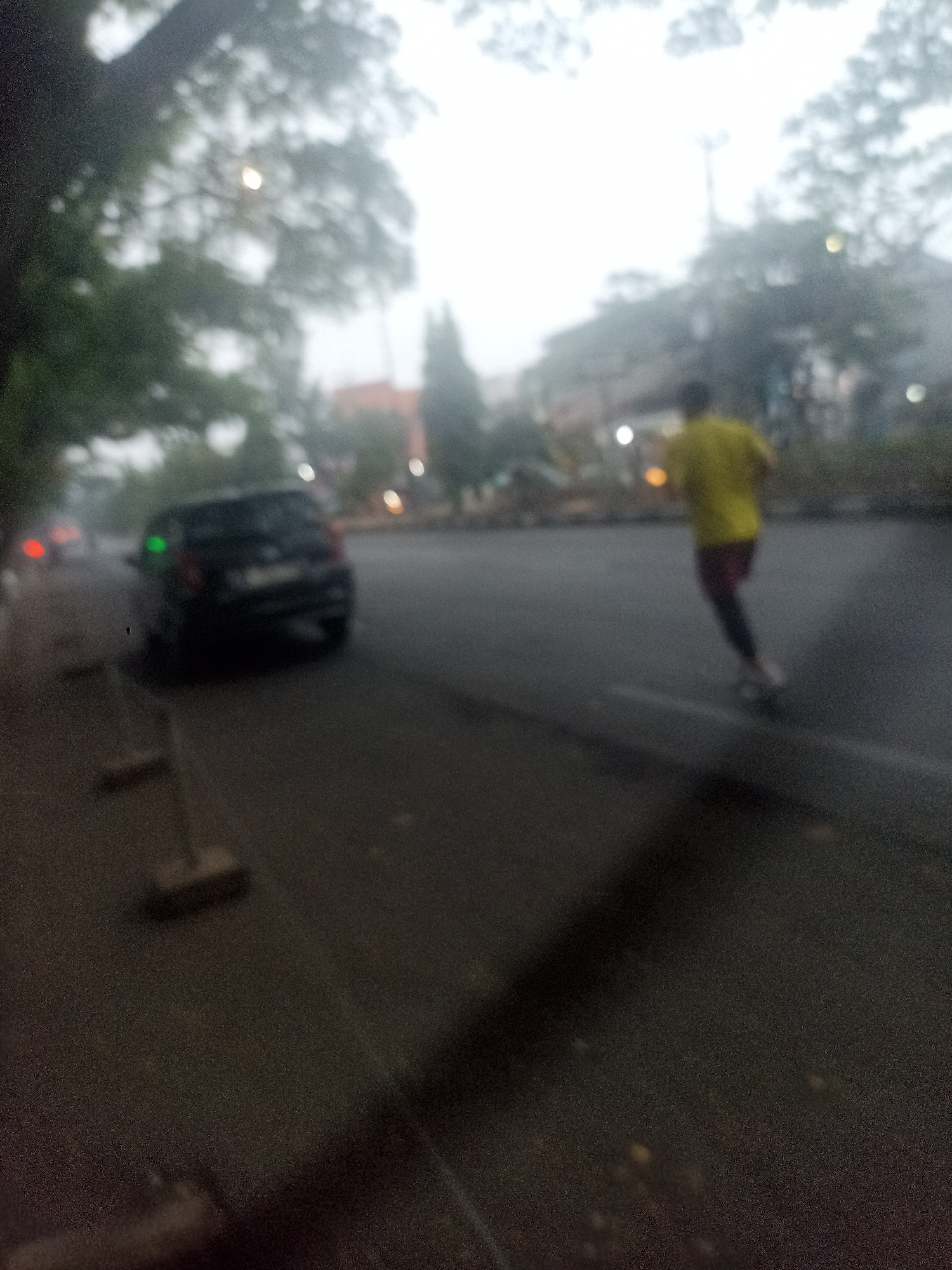 The image captures a slightly blurred street scene, possibly during overcast or foggy weather, suggesting an early morning or late afternoon. In the foreground, a dark-colored car is parked on the left side of the road. Further down the road, a person is seen rollerblading, wearing a bright yellow shirt and dark pants. The road appears to be asphalt. Trees with foliage line the background, and there are indications of buildings or structures in the distance, along with streetlights. The overall ambiance is subdued due to the low light and blurriness.