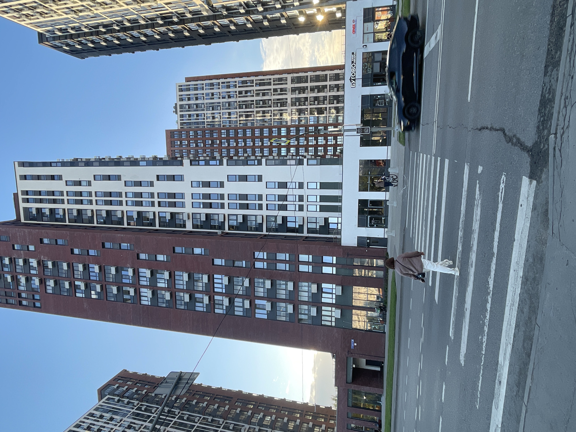 The image shows a street scene in Moscow, Russia, with a focus on modern architecture and urban life. Dominating the frame are several tall, contemporary residential buildings with numerous windows. The buildings vary in facade materials, with one appearing to be brick and another white with dark window frames.
In the foreground, a pedestrian crossing is marked on the asphalt road. A person, dressed in a brown jacket and white pants, is walking across the street on the zebra crossing. They appear to be alone, and their gait suggests a casual pace. A small, light-colored dog on a leash accompanies the person. A black car is visible on the right side of the road, suggesting traffic flow.
The sky is clear and blue, with the sun casting bright highlights on the buildings, indicating it is daytime. The light suggests it might be late afternoon or early morning due to the angled sunlight. The overall atmosphere is calm and orderly, depicting a typical urban environment. Some text is visible on the building on the right, including "БУТОВО" and "ДОМА" which likely relates to the residential area.