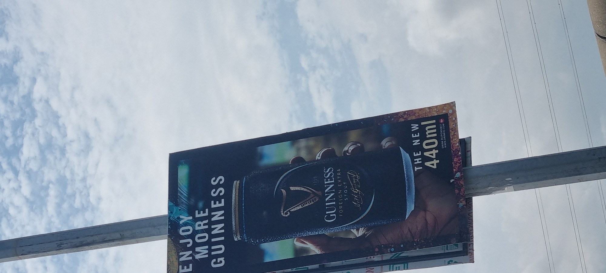 An outdoor Guinness advertisement is displayed on a pole against a cloudy sky. The advertisement prominently features a can of Guinness Foreign Extra Stout held in a hand. The text "ENJOY MORE GUINNESS" is visible at the top of the ad, with "THE NEW 440ml" and the website "DRINKIQ.COM" at the bottom. The advertisement is located on a pole in Ikoyi, Nigeria, with power lines visible in the background.