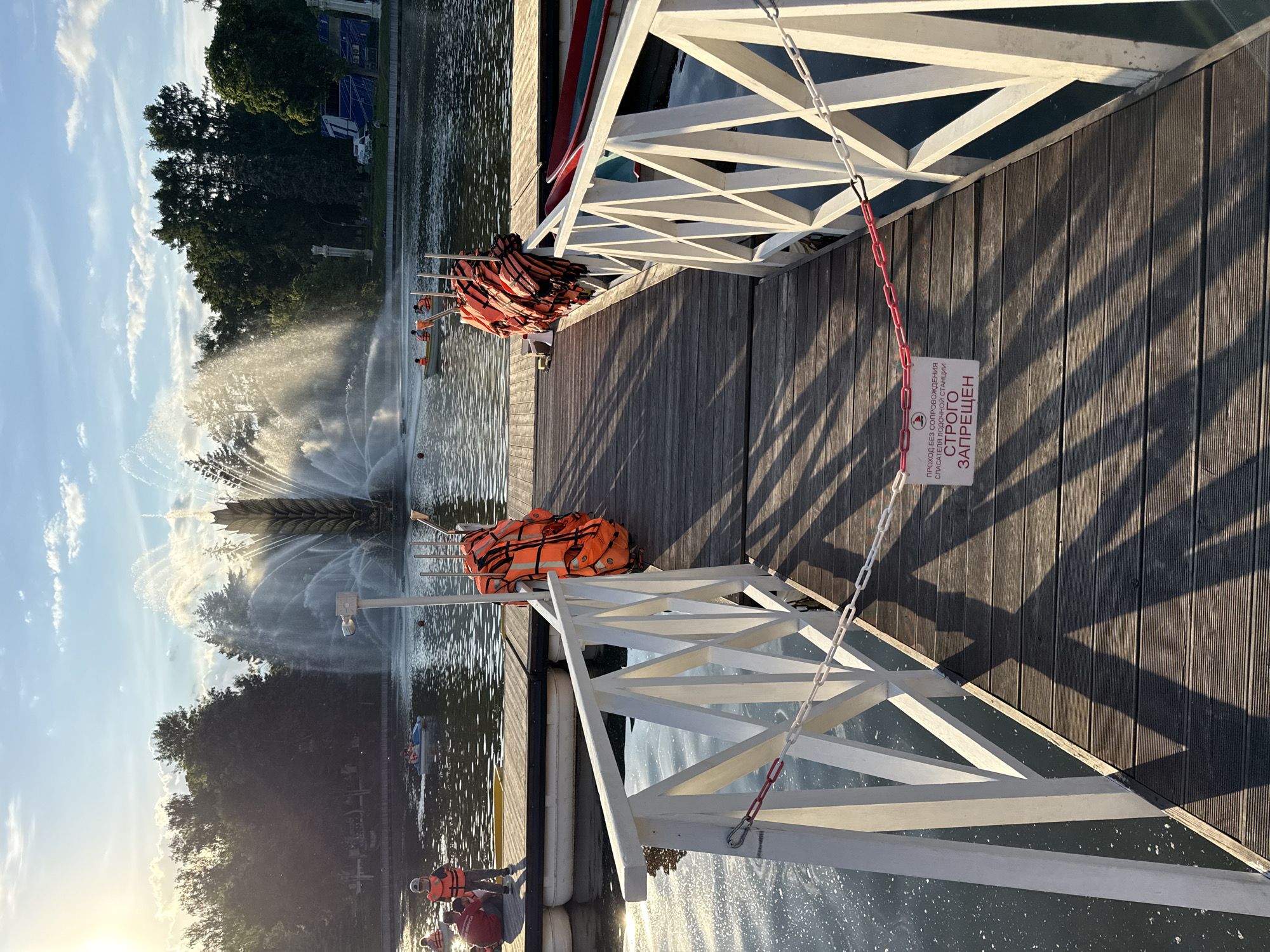 The image captures a sunlit recreational scene at a waterfront area, likely a park, featuring a wooden pier extending into a body of water. In the foreground, a white railing with a geometric pattern borders the pier. A white and red chain hangs from this railing, securing a small white sign with red text. The sign, written in Russian, states: "ПРОХОД БЕЗ СОПРОВОЖДЕНИЯ СПАСАТЕЛЯ ЛОДОЧНОЙ СТАНЦИИ СТРОГО ЗАПРЕЩЕН," which translates to "PASSAGE WITHOUT ACCOMPANIMENT OF THE BOAT STATION RESCUER IS STRICTLY FORBIDDEN."
Numerous bright orange life vests are stacked on poles along the pier and are also visible near the water, indicating water-based activities. Several individuals are present, some wearing life vests, seen on or near small boats and pedal boats further out on the water.
A large, ornate fountain is prominently featured in the mid-ground, actively spraying jets of water into the air. Its mist catches the warm, late afternoon or early evening sunlight, creating a sparkling effect. Long, diagonal shadows are cast across the wooden planks of the pier, signifying the low angle of the sun. The sky is bright blue with scattered white clouds, and lush green trees line the distant shore, completing the tranquil, recreational setting. The specific design of the fountain and the general environment suggest a location within Moscow, Russia.