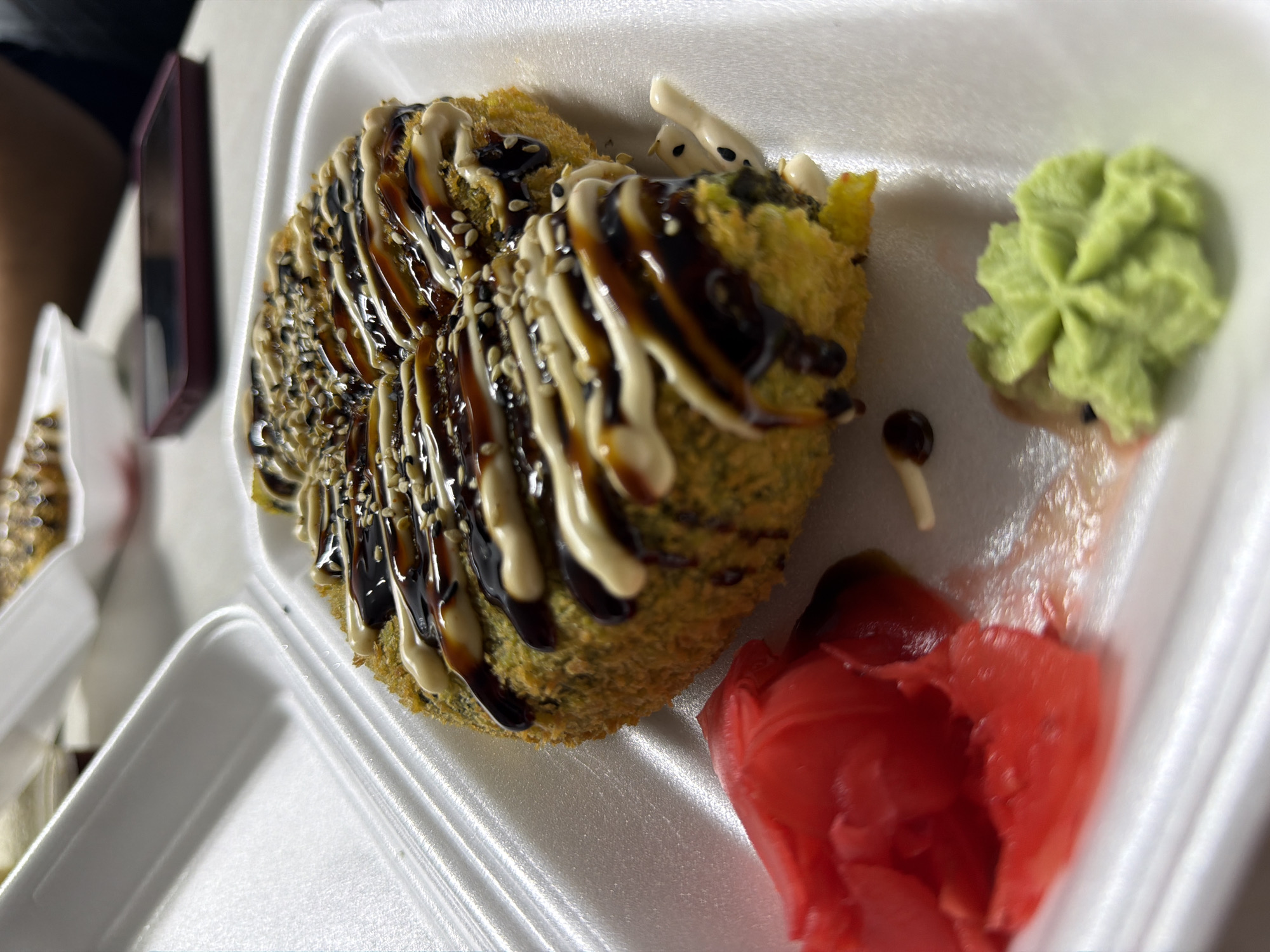 A close-up, low-angle shot shows a deep-fried sushi roll, generously drizzled with a dark sauce and a white creamy sauce. The roll is sprinkled with sesame seeds. To the left of the sushi roll is a pile of pink pickled ginger. To the right, a dollop of bright green wasabi is presented in a decorative swirl. The food is served in a white Styrofoam takeout container, which is positioned on a white surface, possibly a table. In the background, another white takeout container is visible, and a person's hand and arm are partially visible, suggesting the food is being prepared or consumed. The scene appears to be indoors, likely in a restaurant or a home setting in Strășeni, Moldova. The lighting is somewhat dim, with a spotlight effect on the main sushi roll.
