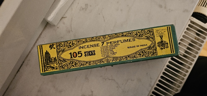 A rectangular box of incense is depicted resting on a light-colored, patterned surface, likely a windowsill or countertop, in an indoor setting within Kamenz - Kamjenc, Germany. The box is oriented diagonally, with its long side extending from the lower left to the upper right of the frame. It features a predominantly yellow face with a green border and black text and illustrations.
The text on the box reads "105 STICKS" vertically on the left, and "INCENSE 7 PERFUMES" horizontally on the right. Below "INCENSE 7 PERFUMES," a partial list of perfumes is visible, including "7 STICKS TULSY-AMBER," "15 STICKS SANDAL," "15 STICKS ROSE," "15 STICKS CHAMPA," "15 STICKS RAJNEEH," "15 STICKS JASMINE," and "15 STICKS LILAC." The phrase "MADE IN INDIA" is printed vertically near the center.
Illustrations include a dark silhouette of a building, possibly a temple or dome, with a floral emblem at the top of the box, and a stylized tree in a landscape at the bottom. The background behind the box consists of a plain light-colored wall. In the lower left of the image, a white, metal heating radiator with visible grates and some dust accumulation runs horizontally. No individuals are present in the scene.