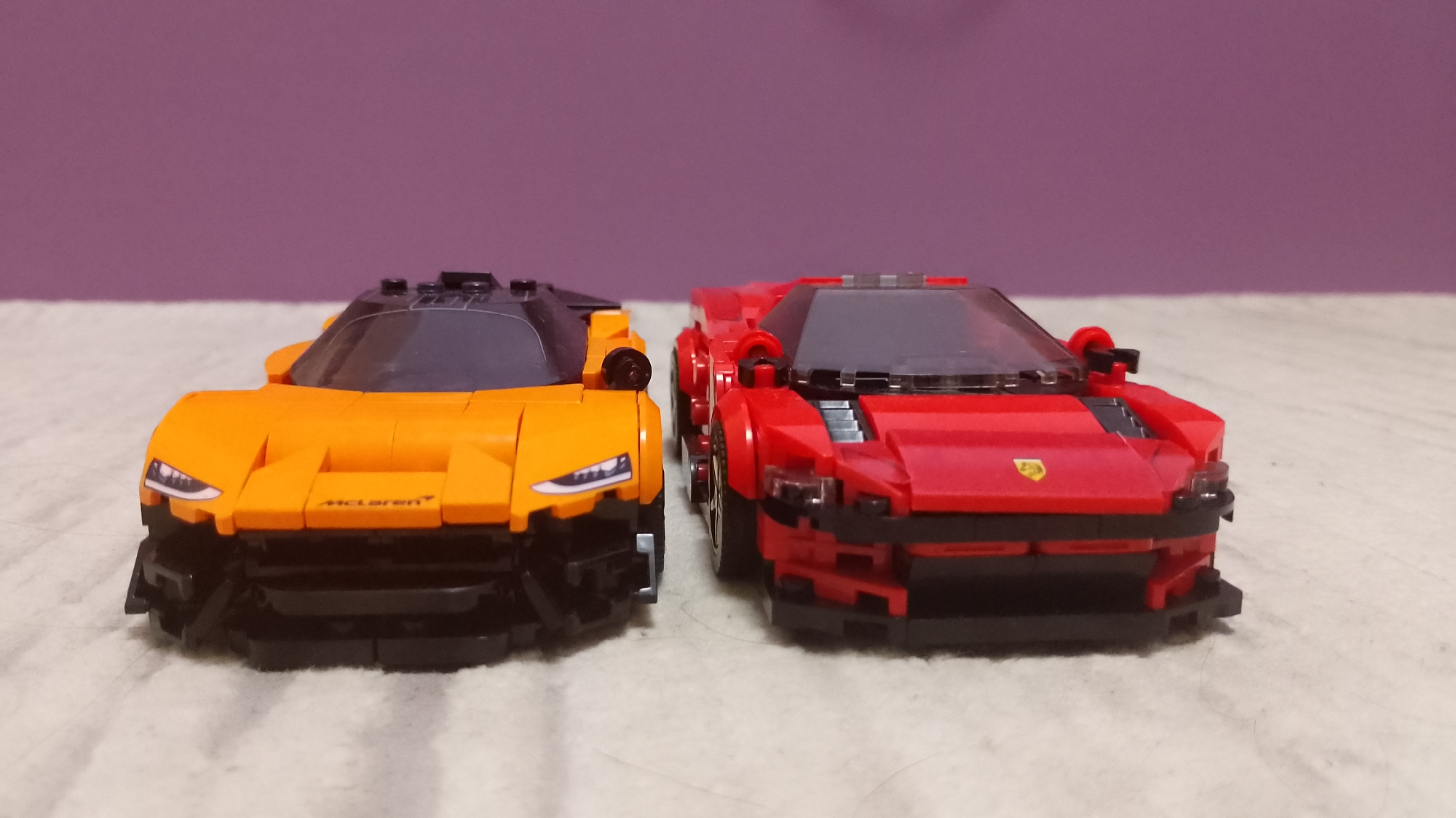 The image displays two Lego toy cars positioned side-by-side on a light-colored, textured surface. Behind them is a blurred, plain purple background.
The car on the left is predominantly orange with black accents for the roof, windshield, and lower front bumper. White text, "McLaren," is visible on its front hood, confirming its model. The car features detailed headlights.
The car on the right is primarily red with black components for its roof, windshield, and lower front. A small yellow shield-shaped logo, characteristic of Ferrari, is affixed to its front hood.
Both cars are low-profile sports car models, facing forward towards the viewer. The orange McLaren is slightly closer to the camera and positioned to the left of the red Ferrari. The scene appears to be indoors, under even, bright lighting.