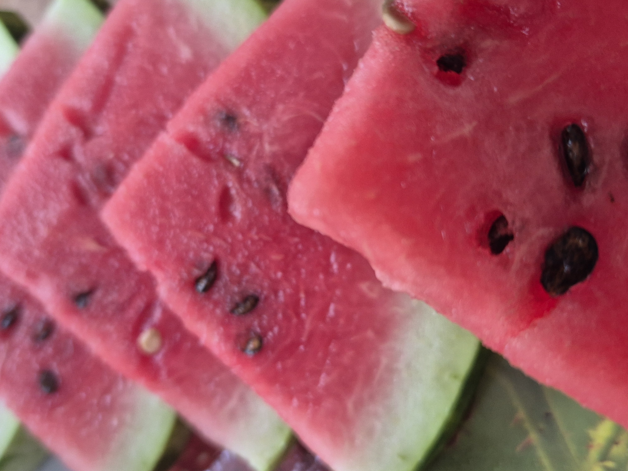 The image is a close-up, overhead view of several slices of watermelon arranged in a staggered fashion. The watermelon slices are vibrant pink with visible seeds. The rind, a gradient of green to white, is visible on the edges of some slices. The lighting is natural and bright, highlighting the juicy texture of the fruit. There is no discernible urban or natural environment, as the focus is entirely on the watermelon. The scene does not depict any specific location in Kokshetau, Kazakhstan, or any other city.