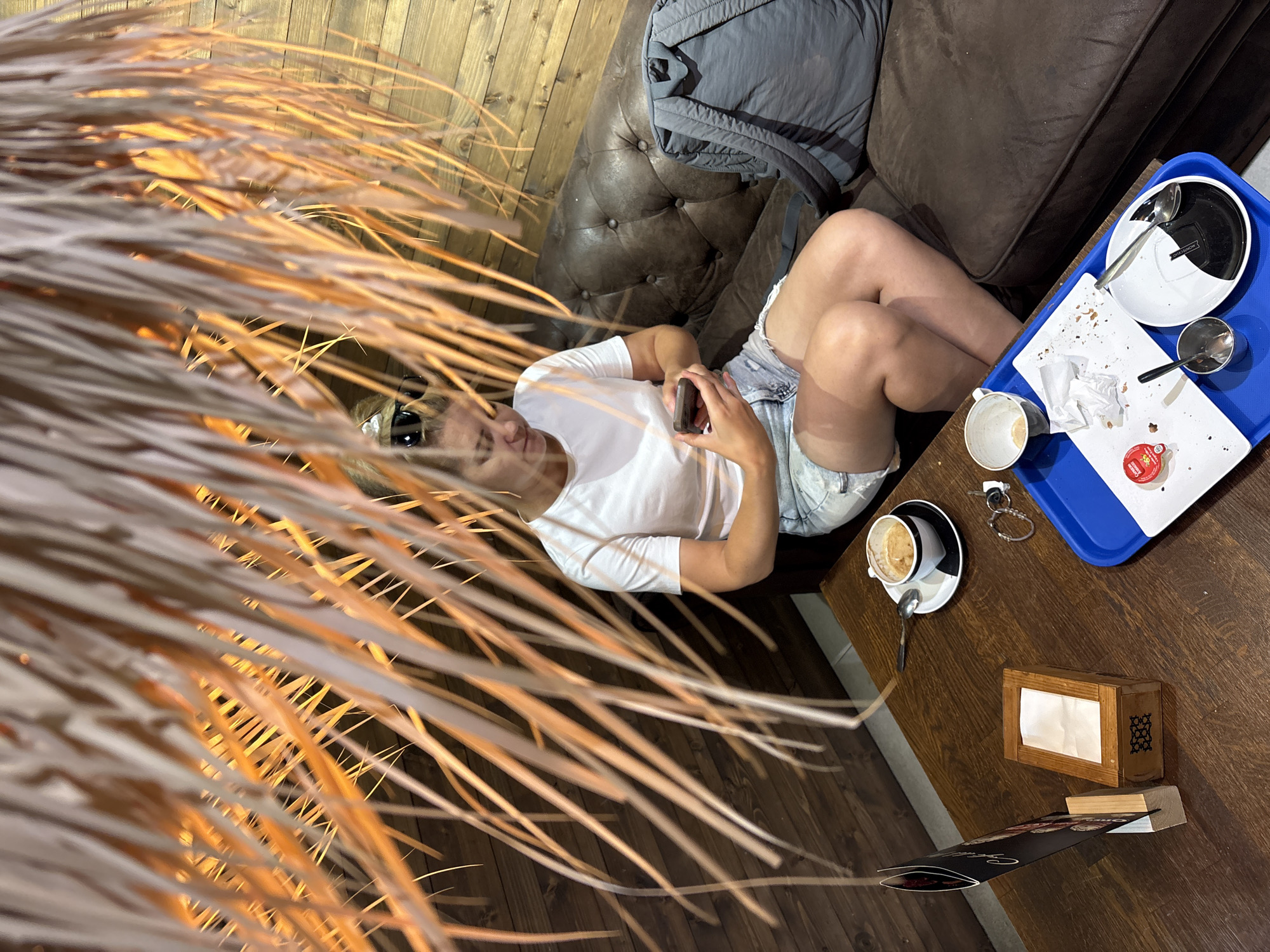 The image captures a relaxed scene, likely in a cafe or lounge, with a person lounging on a dark brown leather sofa. The person, wearing a white t-shirt and denim shorts, is holding a phone and appears to be engrossed in it. A grey backpack is placed on the sofa behind them. In front of the sofa, on a wooden table, is a blue tray containing remnants of a meal, including a plate with crumbs, a used napkin, an empty coffee cup on a saucer, a small red packet, and a spoon. Another coffee cup, also with a used coffee, sits on a saucer beside keys and a small metal object. A wooden napkin holder with white napkins is also visible on the table, along with a menu or flyer placed at an angle. The background features a wooden-paneled wall. The lighting suggests it could be daytime or evening, with artificial light casting a warm glow. The overall atmosphere is casual and comfortable, with the dry, feathery plant fronds in the foreground adding a natural, perhaps slightly bohemian, touch to the scene.
